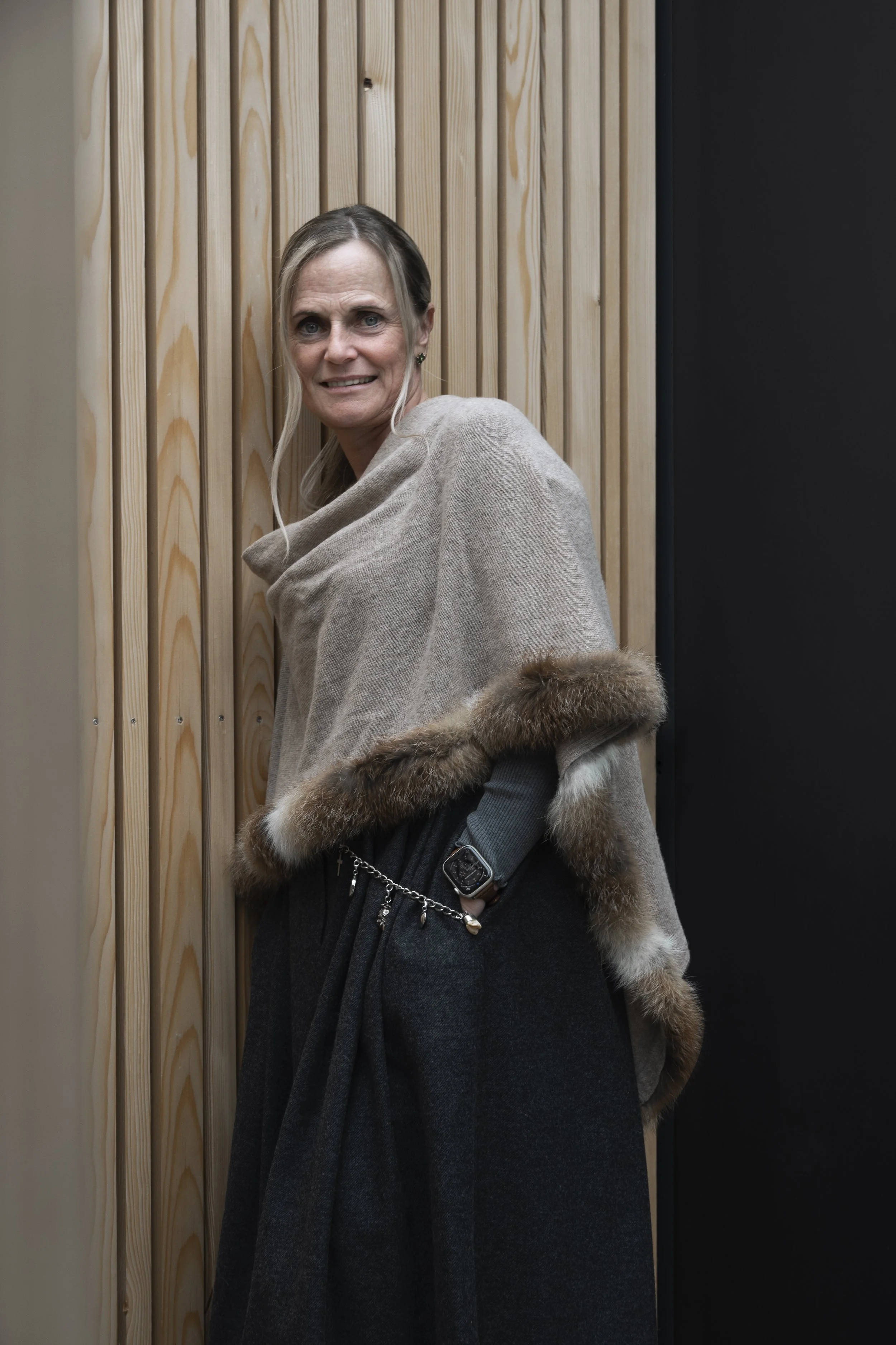 Eine Frau steht vor einer Wand aus Holzlatten, trägt einen grauen Mantel mit Fellbesatz an den Ärmeln und schaut in die Kamera.