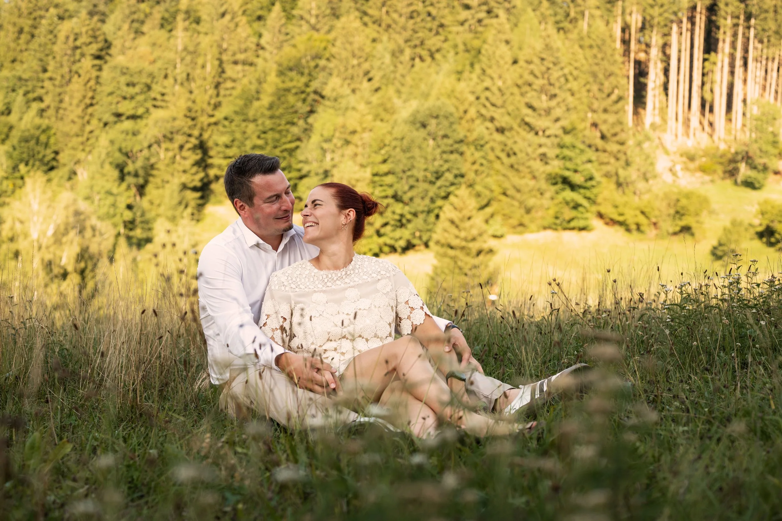 Ein Paar sitzt hintereinander in der Wiese und blickt sich lächelnd in die Augen.