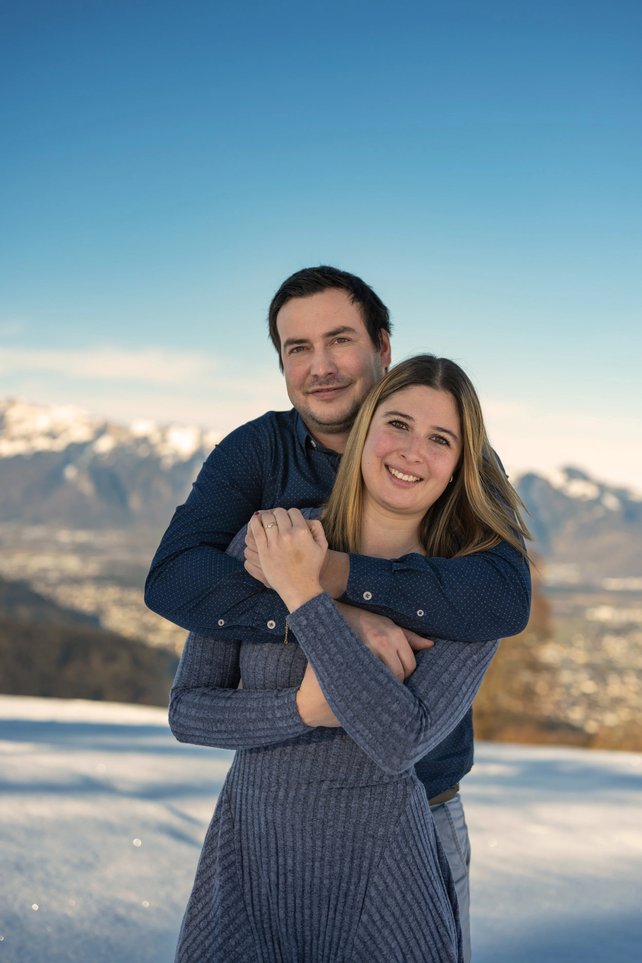Paar umarmt sich liebevoll und blickt lächelnd in die Kamera. Im Hintergrund schneebedeckte Berge.