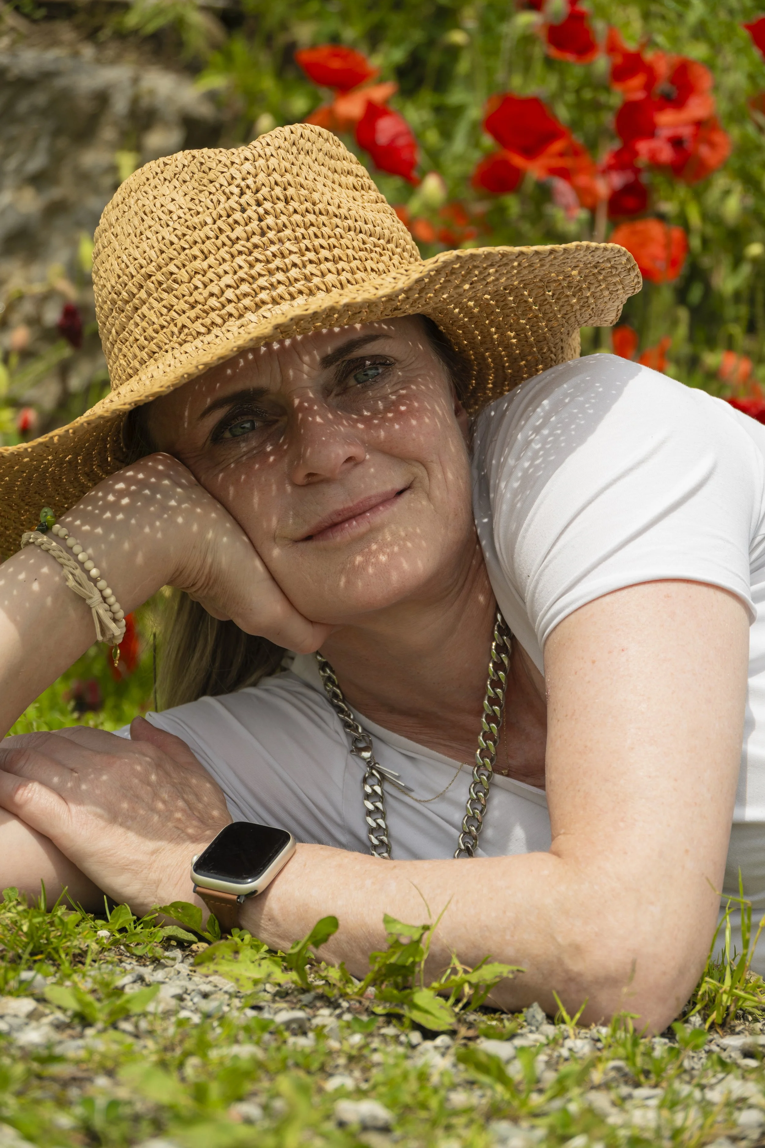 Eine lächelnde Frau liegt auf dem Boden, trägt einen Strohhut, ein weißes T-Shirt, eine Kette und eine Smartwatch, umgeben von roten Blumen im Hintergrund.
