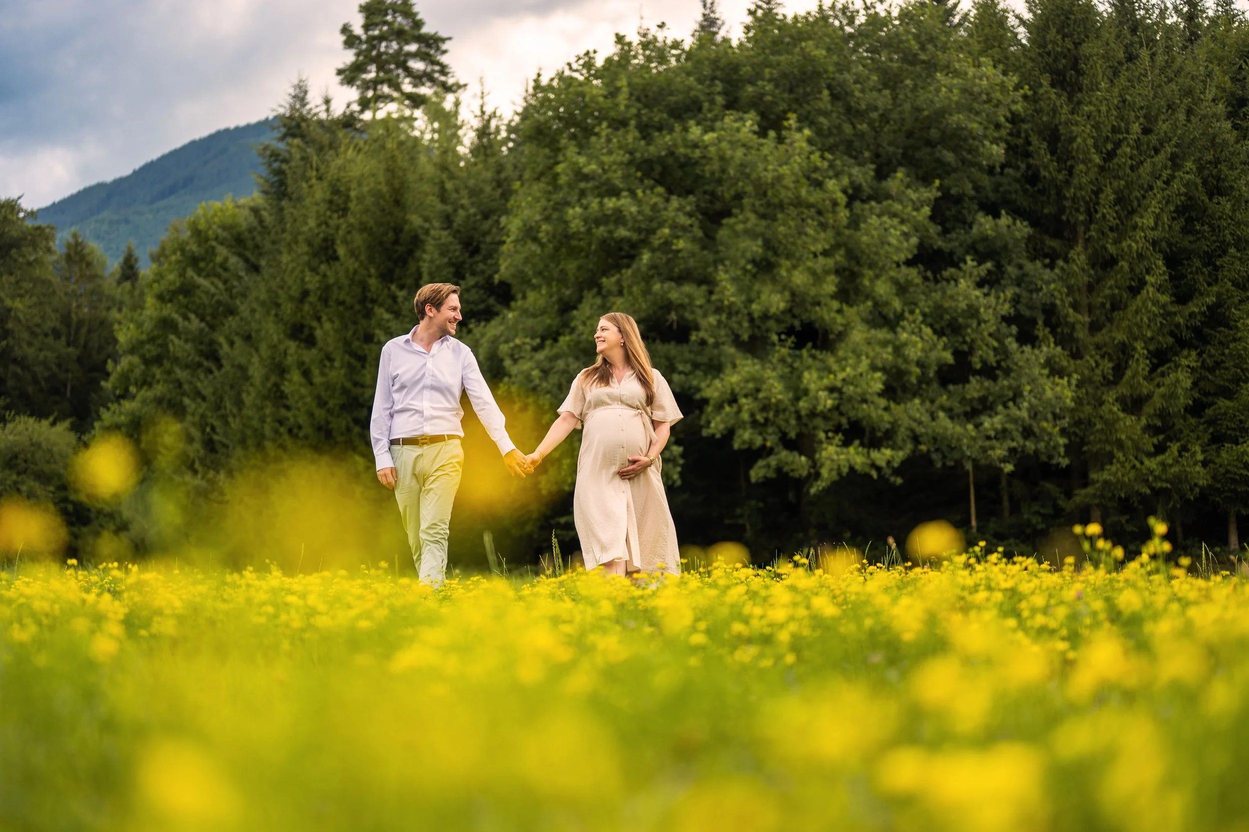 Ein Paar hält sich an der Hand und lächelt sich an, während die Frau ihren wunderschönen Babybauch streichelt. Sie gehen gemeinsam über eine Wieso voller gelber Blumen.