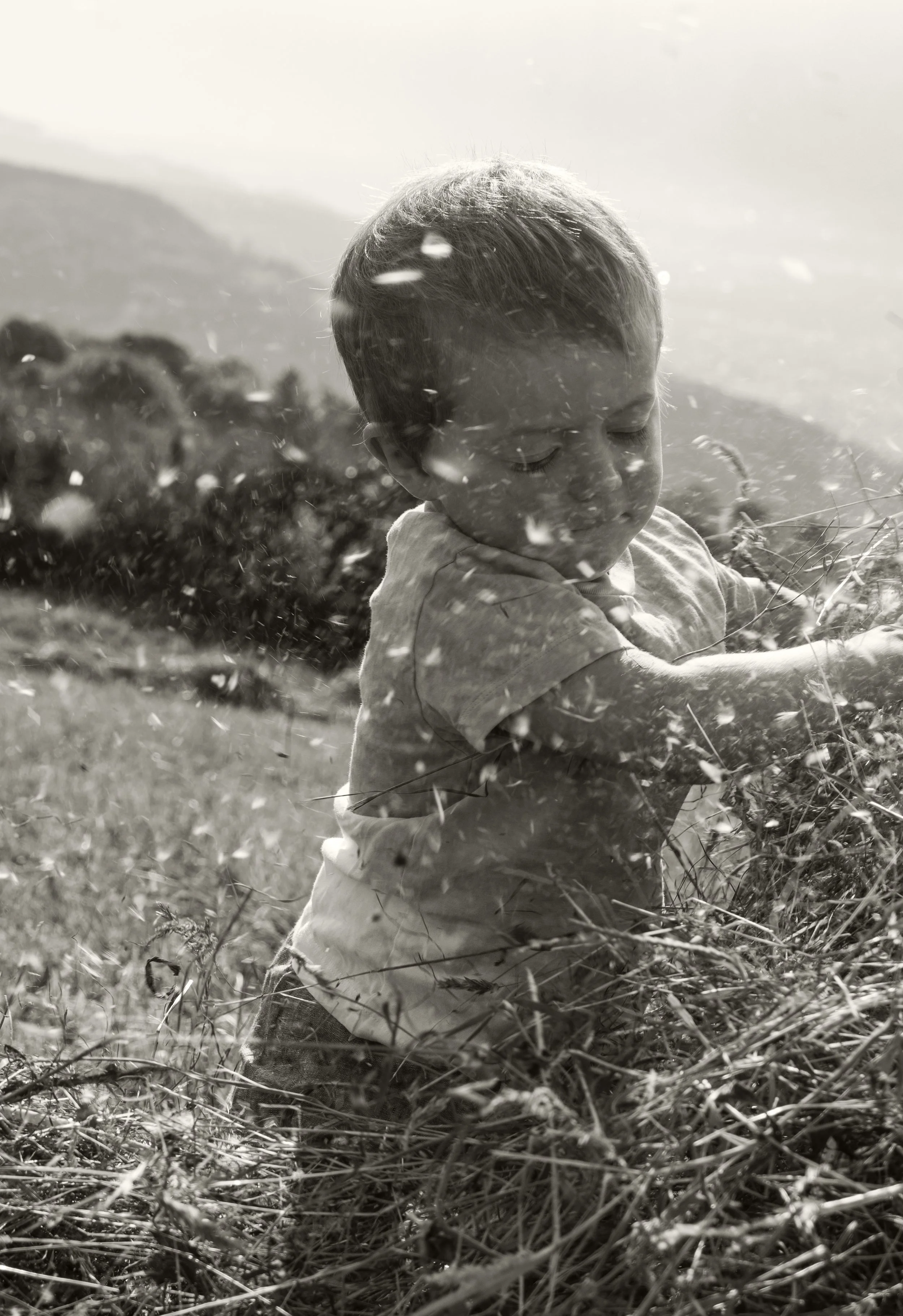 Ein Junge spielt im Freien mit trockenem Gras, in Schwarz-Weiß aufgenommen.