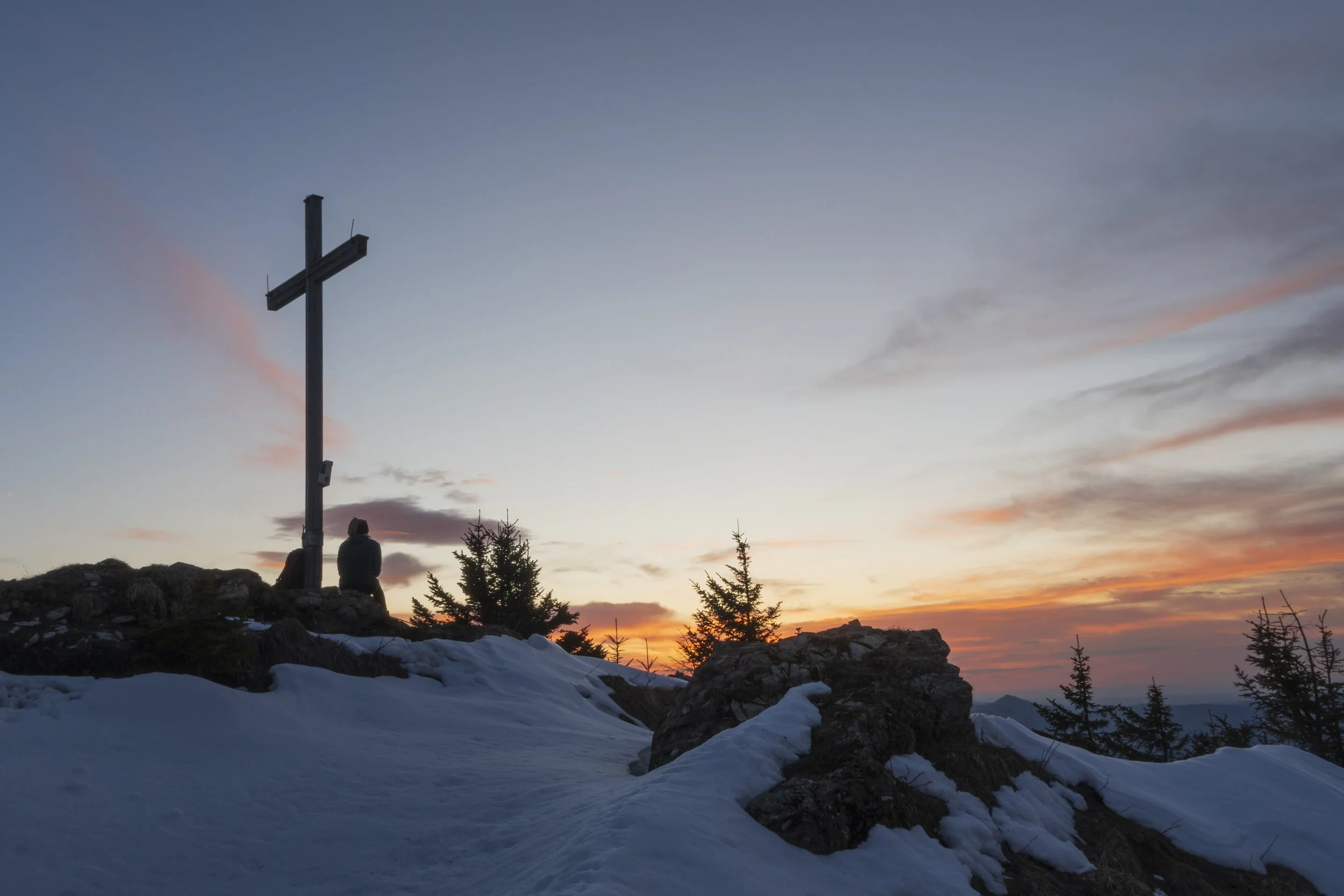 Sonnenuntergang auf einem Berggipfel, mit Schnee bedecktem Boden, einem großen Holzkreuz und einer Person, die am Gipfel sitzt.