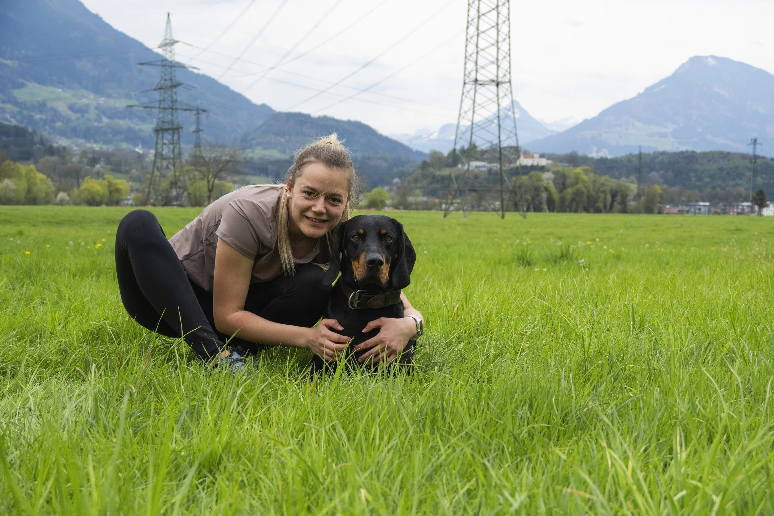 Eine junge Frau sitzt tief unten in einer grünen Wiese, hält einen Hund fest und lächelt, im Hintergrund Berge und Hochspannungsmasten.