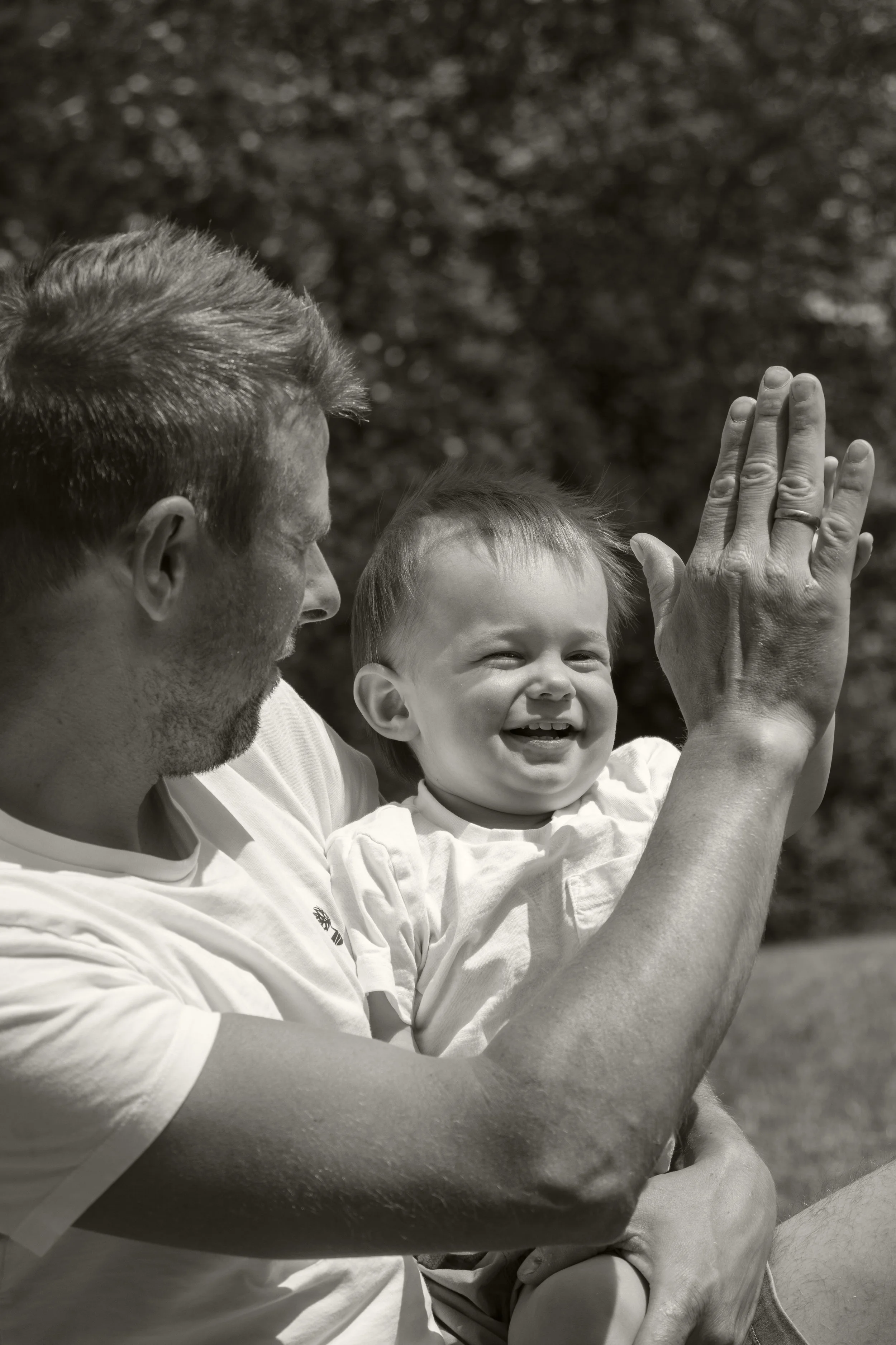 Ein Mann hält ein lachendes Kleinkind in den Armen, beide sitzen draußen, im Hintergrund sind Bäume, das Bild ist schwarz-weiß.