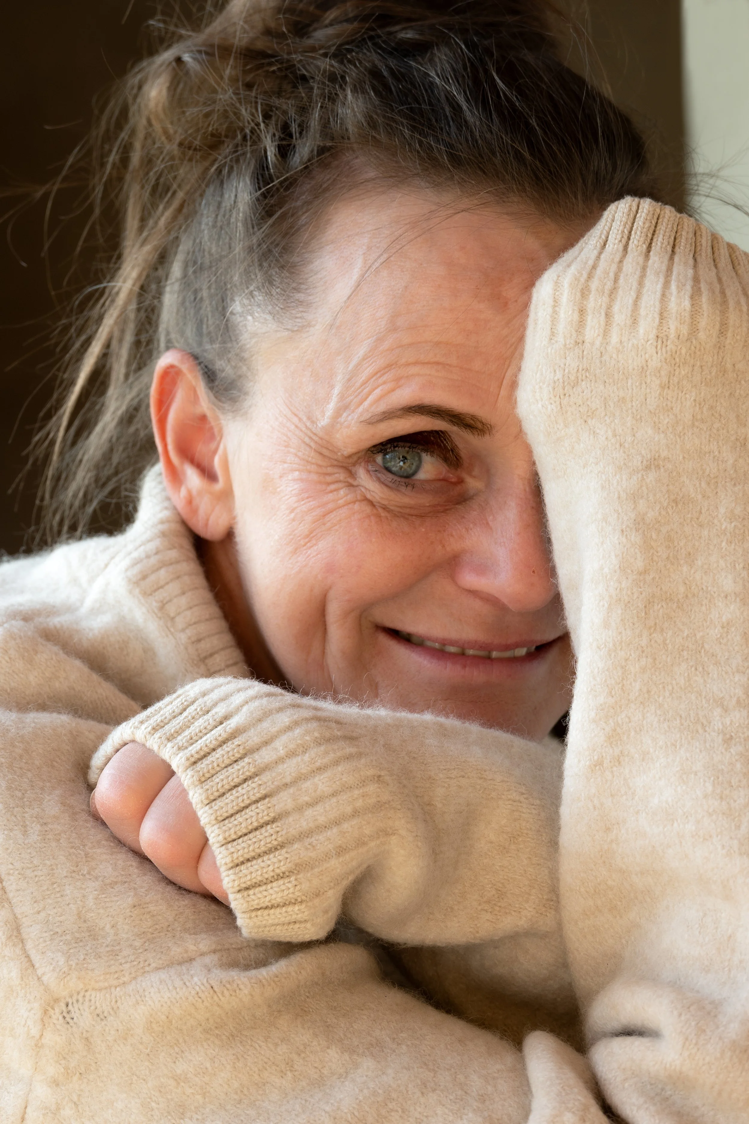 Nahaufnahme einer lachenden Frau mit grau-blauen Augen, die einen cremefarbenen Strickpullover trägt und sich die Hand gegen die Stirn hält.
