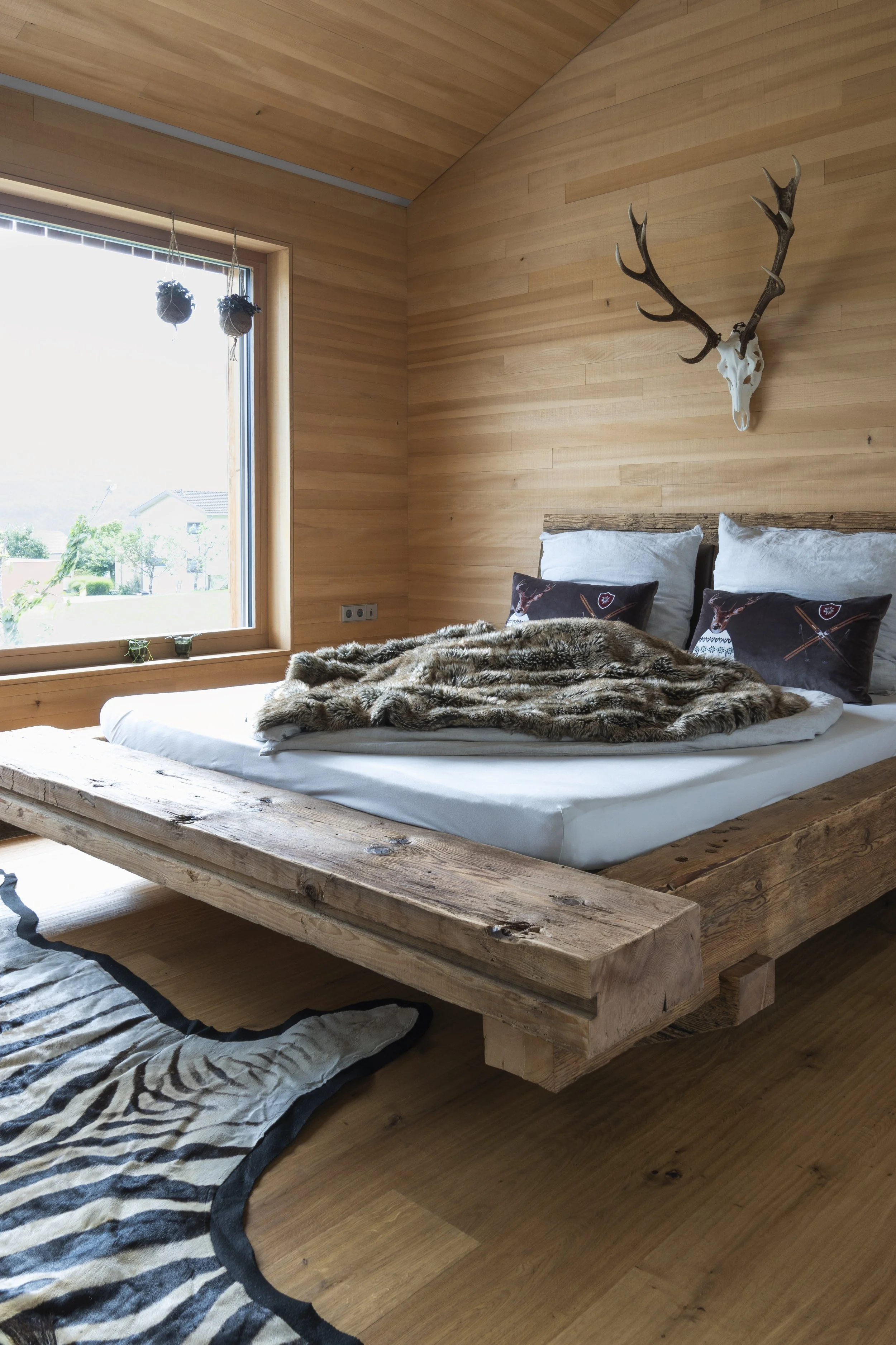 Ein Bett mit rustikalem Holzrahmen, kuscheliger Felldecke und Schlafkissen. An der Wand über dem Bett hängt ein Hirschgeweih. Den Fußboden ziert ein schönes Zebrafell.