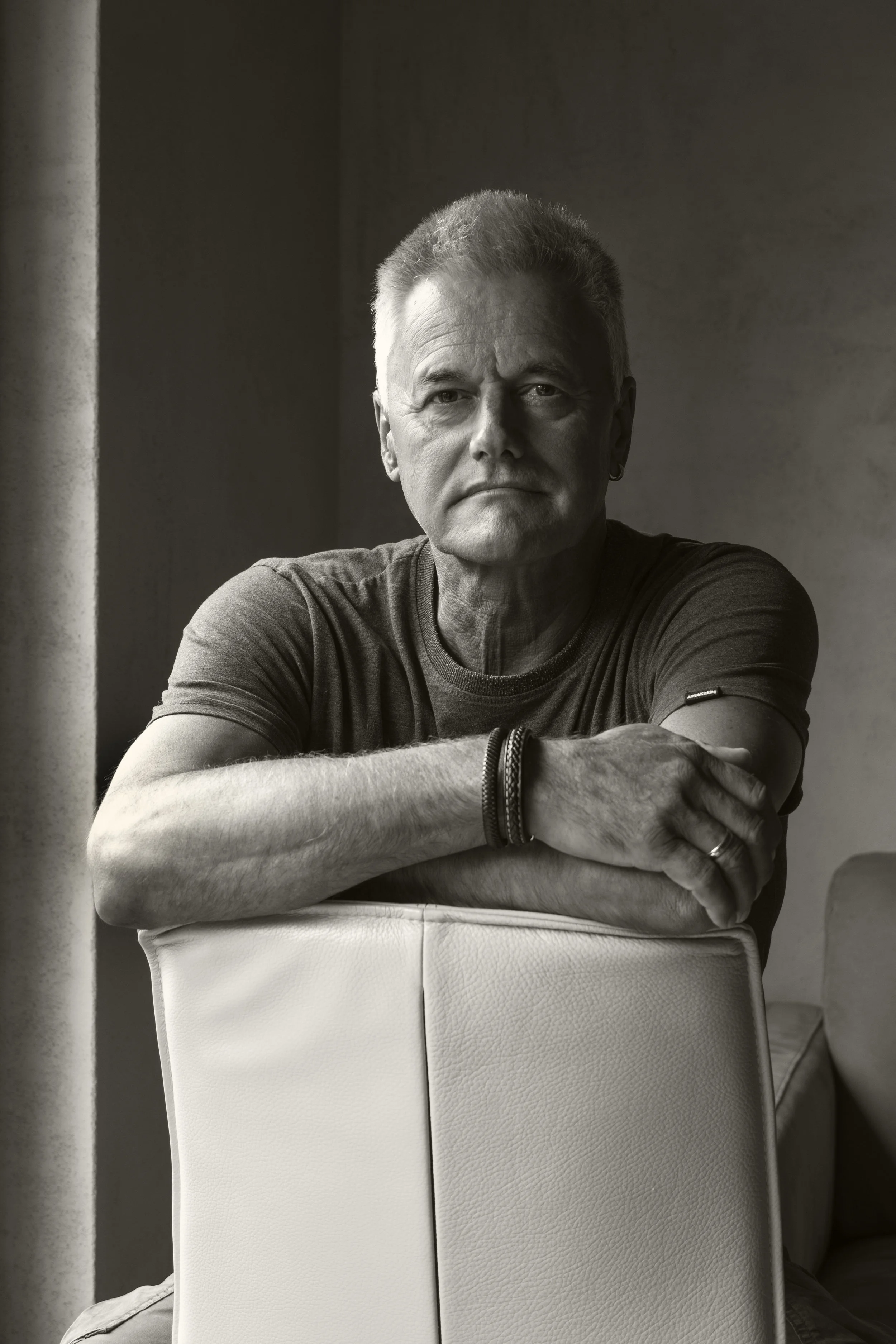 Schwarzweiße Porträtfotografie eines älteren Mannes, der nach vorne schaut, auf einem weißen Stuhl sitzt, mit einem Arm auf der Rückenlehne, trägt ein dunkles T-Shirt, trägt Armbänder an einem Handgelenk, in einem ruhigen Raum mit dunklem Hintergrund