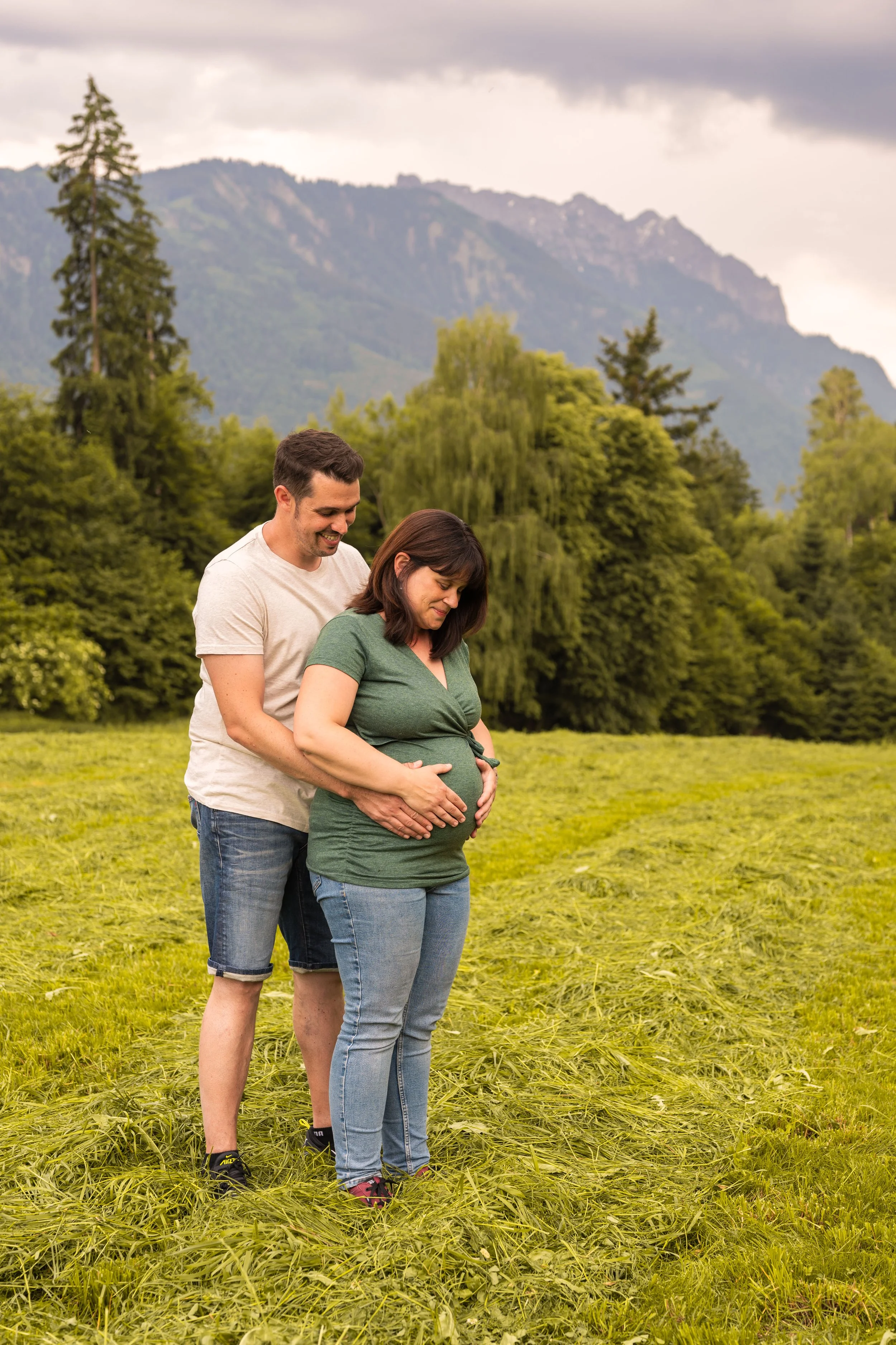 Ein Paar steht hintereinander, sie halten gemeinsam den Babybauch der Frau und blicken fröhlich nach unten.