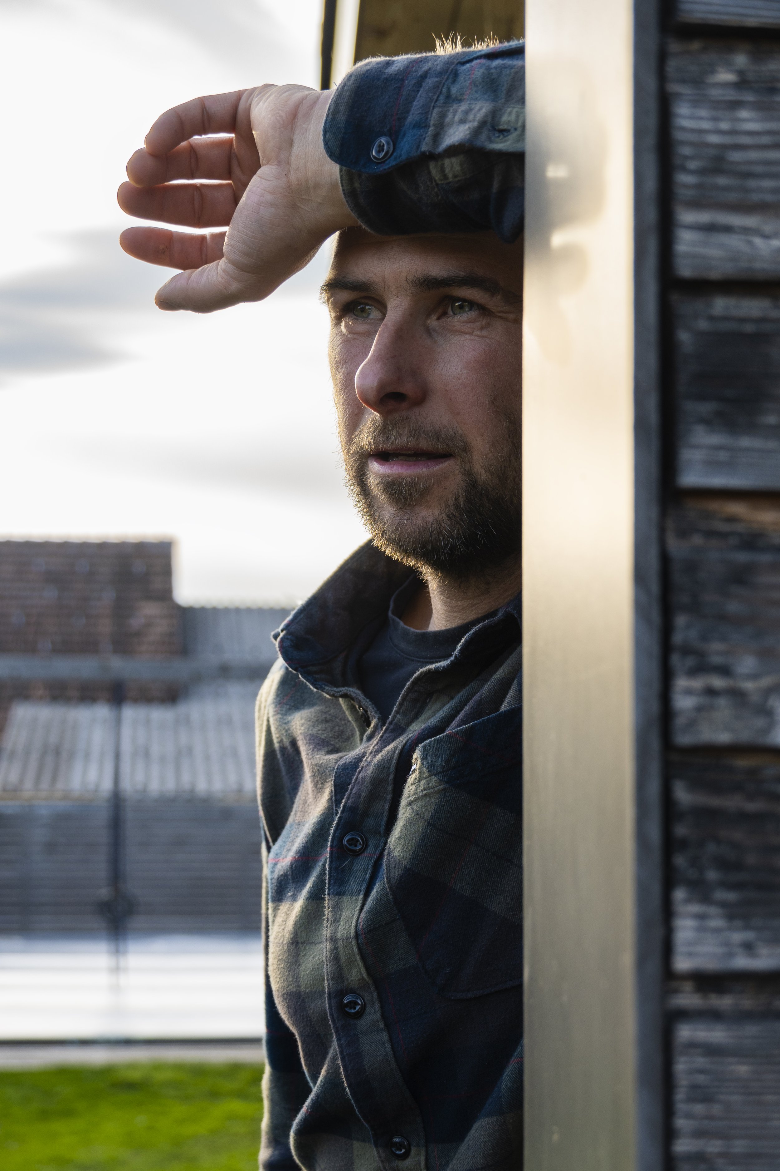 Mann blickt nach draußen, lehnte an einer Wand, trägt Hemd mit Flanellmuster, Sonnenlicht im Hintergrund.