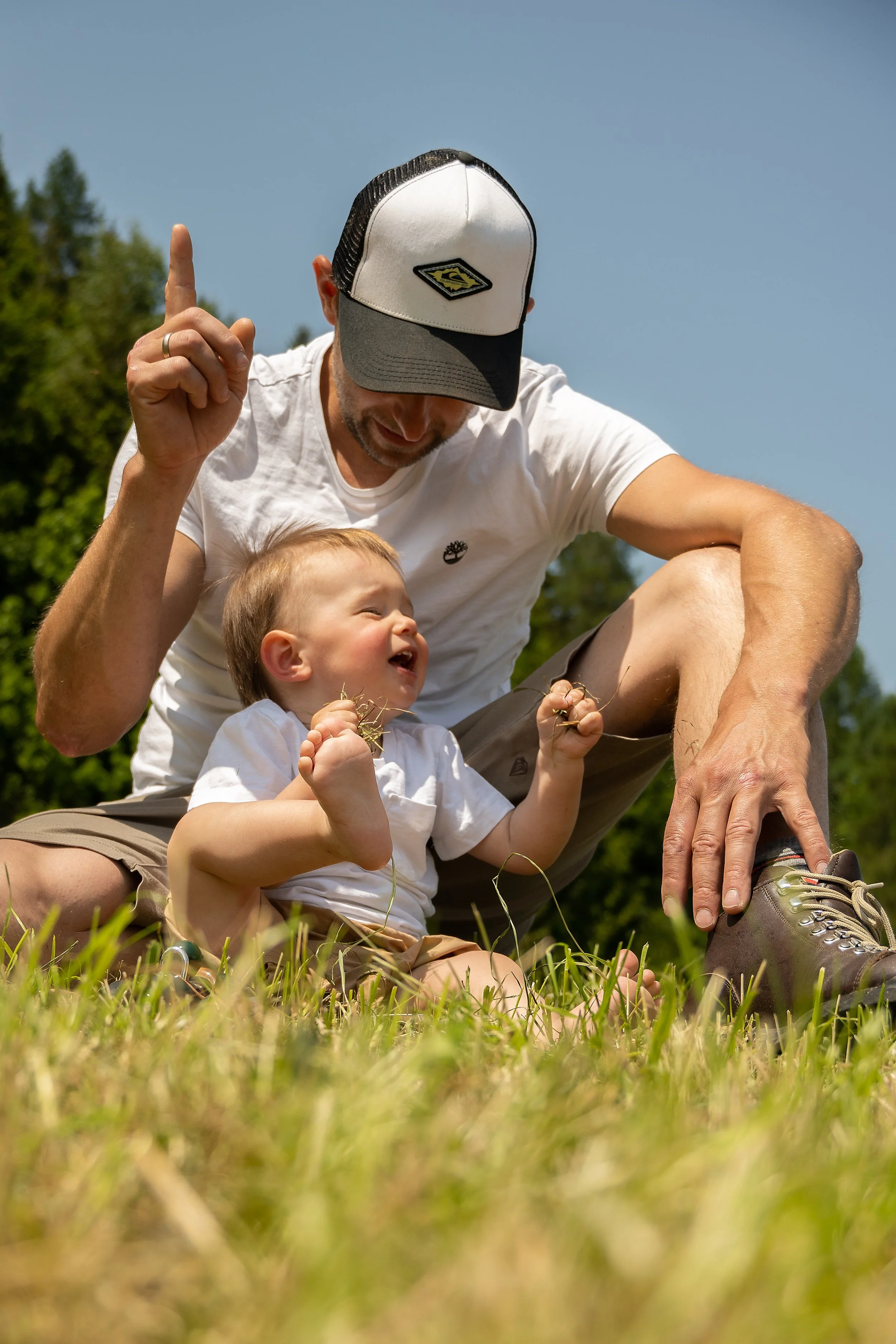 Ein Mann mit einem Kind, die im Freien auf dem Gras sitzen, lachen und spielen, umgeben von Bäumen an einem sonnigen Tag.