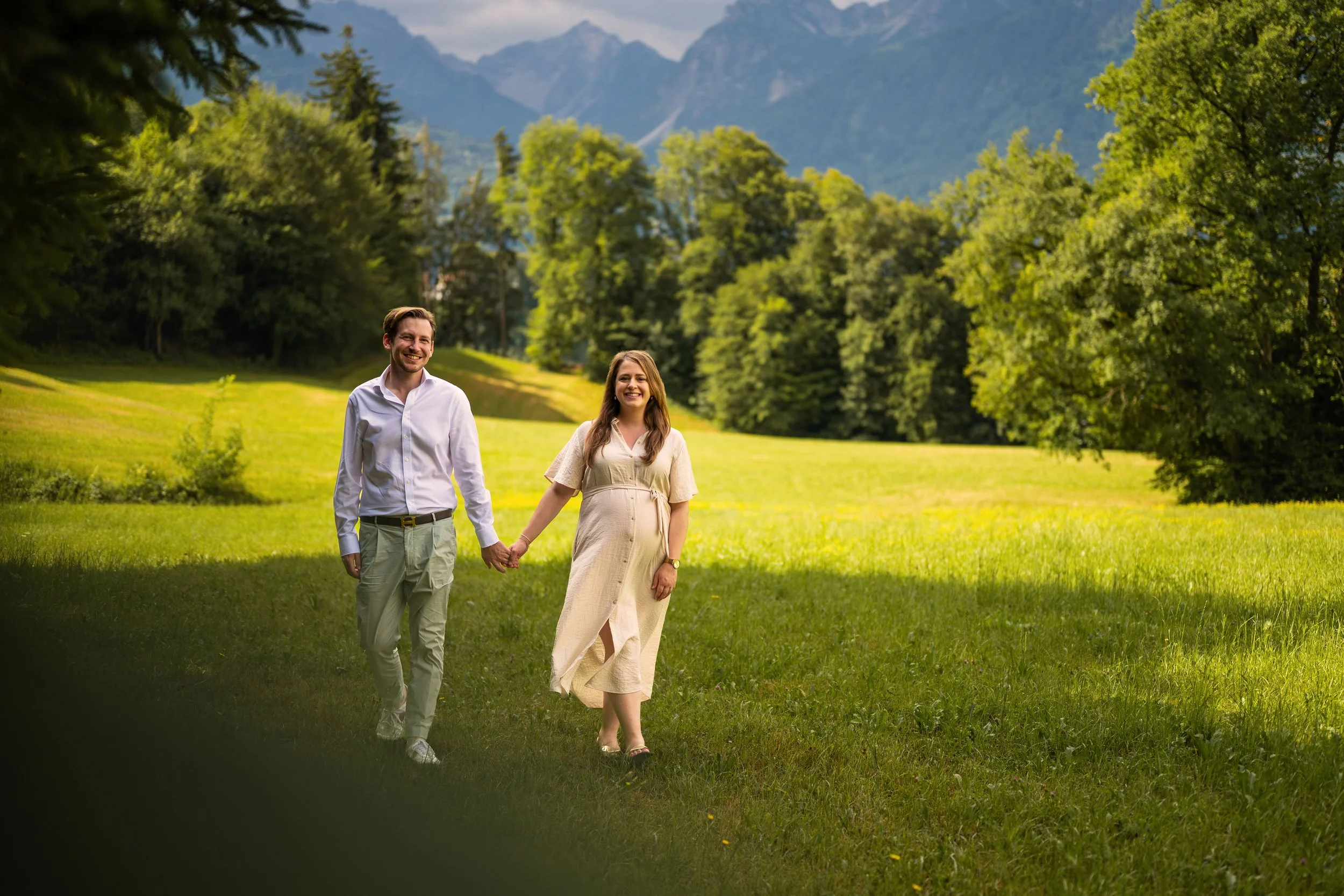 Ein Paar hält sich an der Hand und streift lächelnd über eine saftig grüne Wiese. Im Hintergrund eine schöne Bergkulisse.