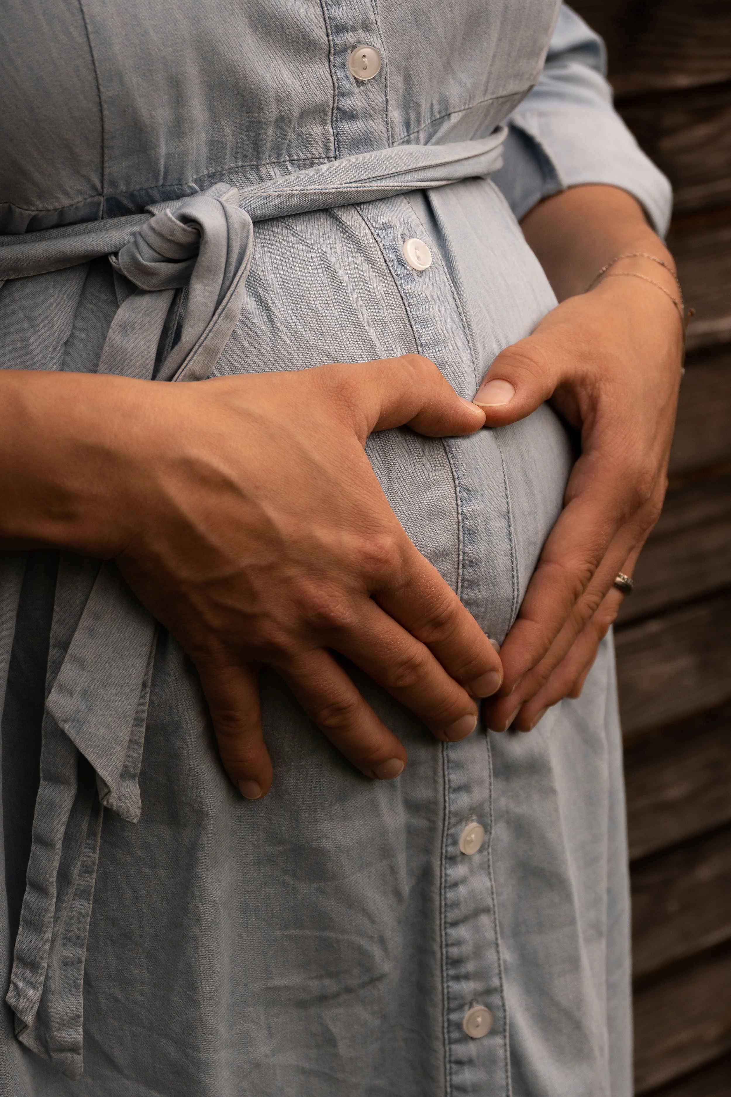 Eine schwangere Person trägt ein hellblaues Kleid mit Knöpfen und einer Schleife in der Taille. Zwei Hände formen ein Herz auf ihrem Babybauch.