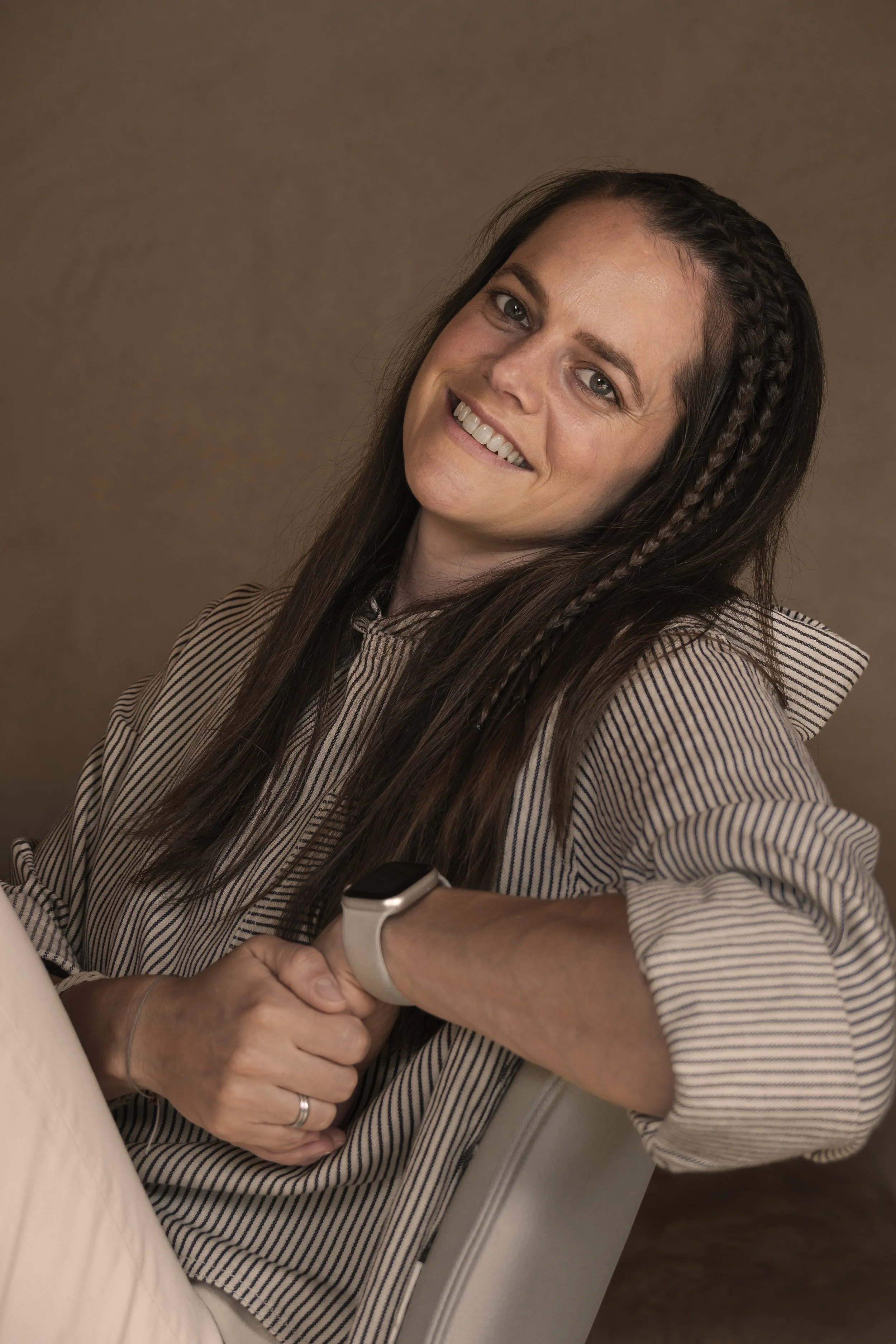 Frau mit braunen, geflochtenen Haaren, lächelnd, sitzt an einem Tisch, trägt eine Armbanduhr, schaut in die Kamera.