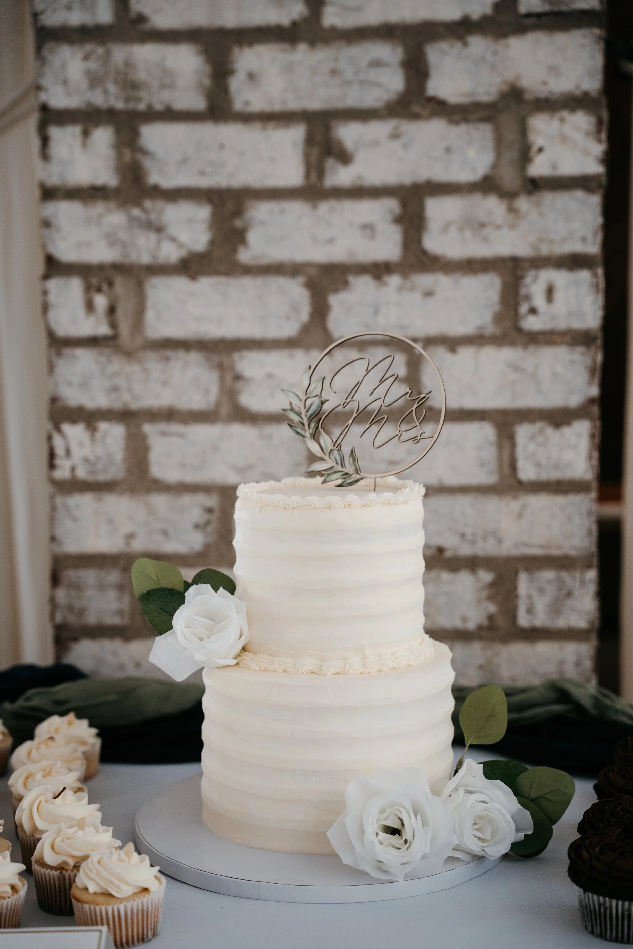 Two-tier white wedding cake with floral decorations and a gold "Mr & Mrs" cake topper, decorated with white roses and green leaves, placed on a silver cake board.