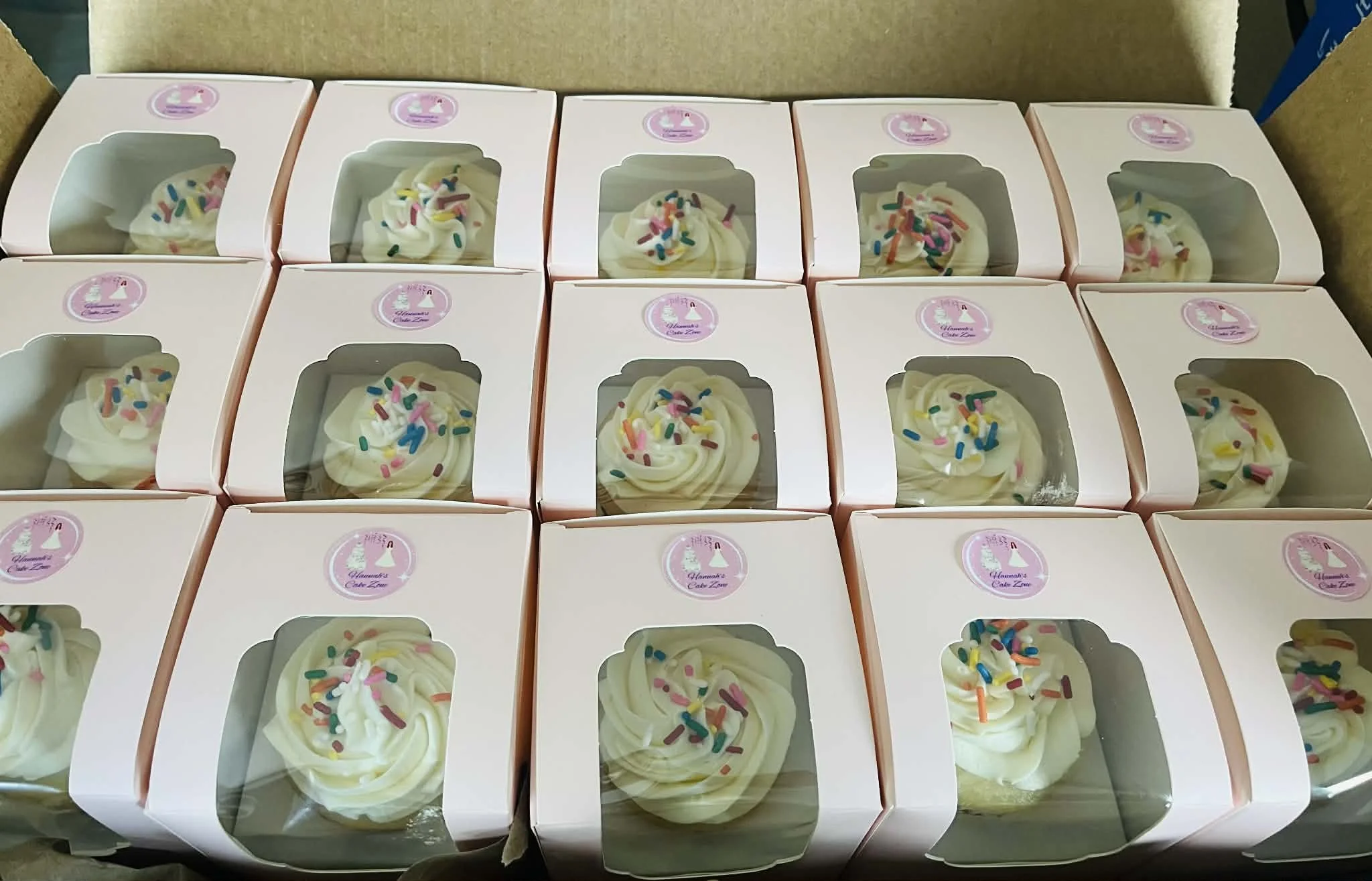 Individually packaged cupcakes with white frosting and colorful sprinkles in pink boxes with clear windows.