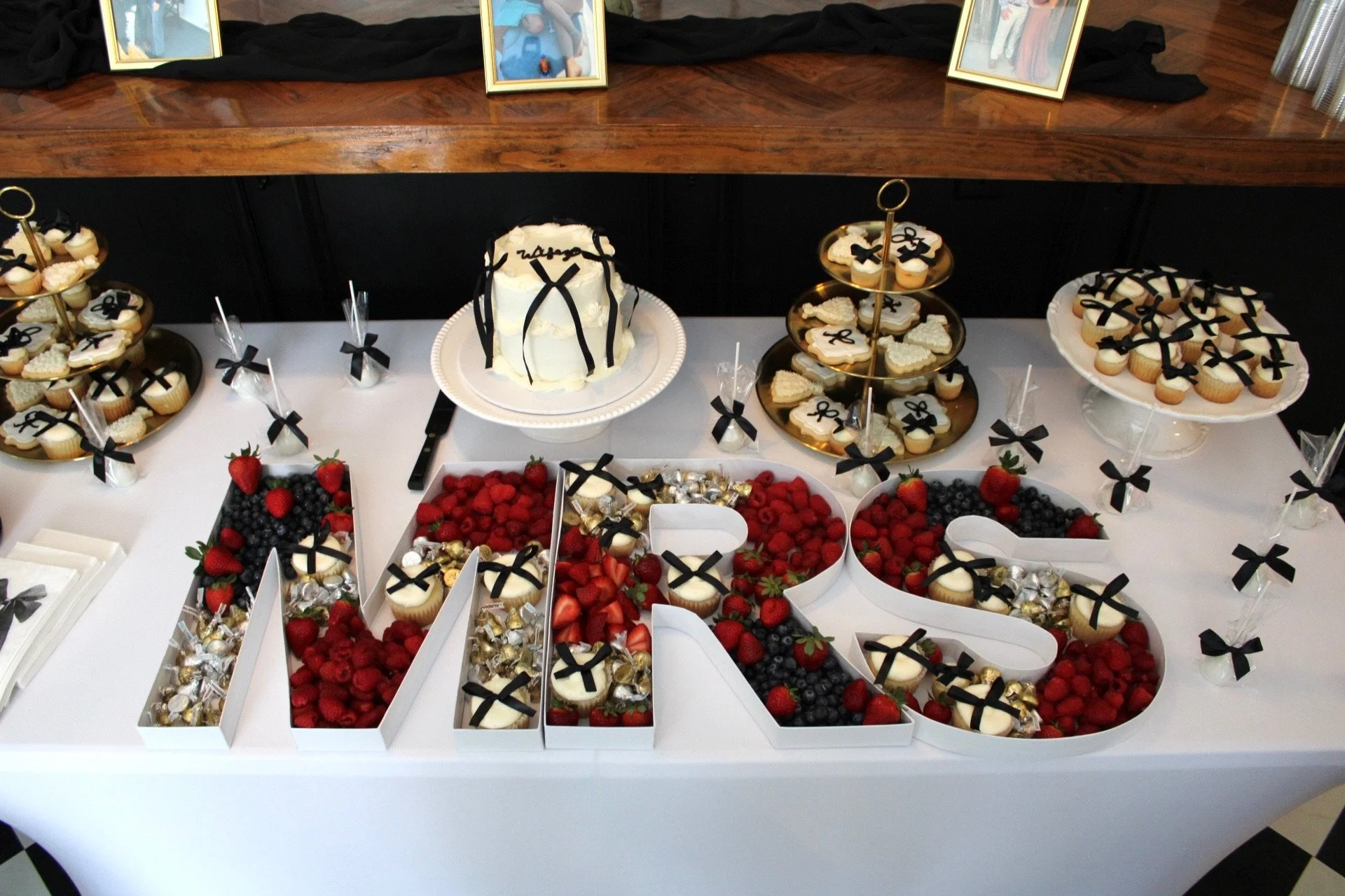 A dessert table featuring a white and black bow theme, cake, cupcakes, cookies, and cake pops with black ribbon toppings, and a large sign spelling 'MR & MRS' filled with various berries and chocolates, all arranged on white and gold platters.