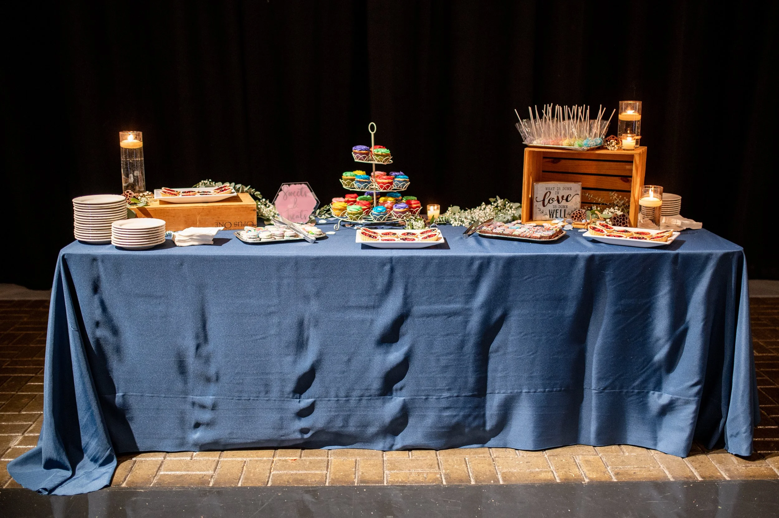 Dessert table with blue tablecloth, featuring stacking macarons, cupcakes, and cookies, surrounded by candles, plates, and decorative elements.