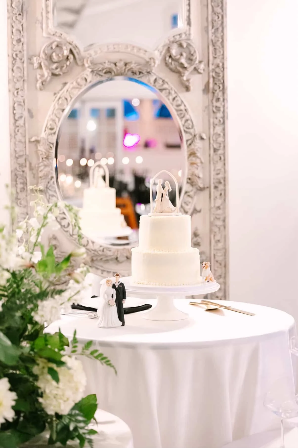 A wedding cake with a bride and groom topper, placed on a white table with a floral arrangement, in front of a vintage mirror with ornate frames.