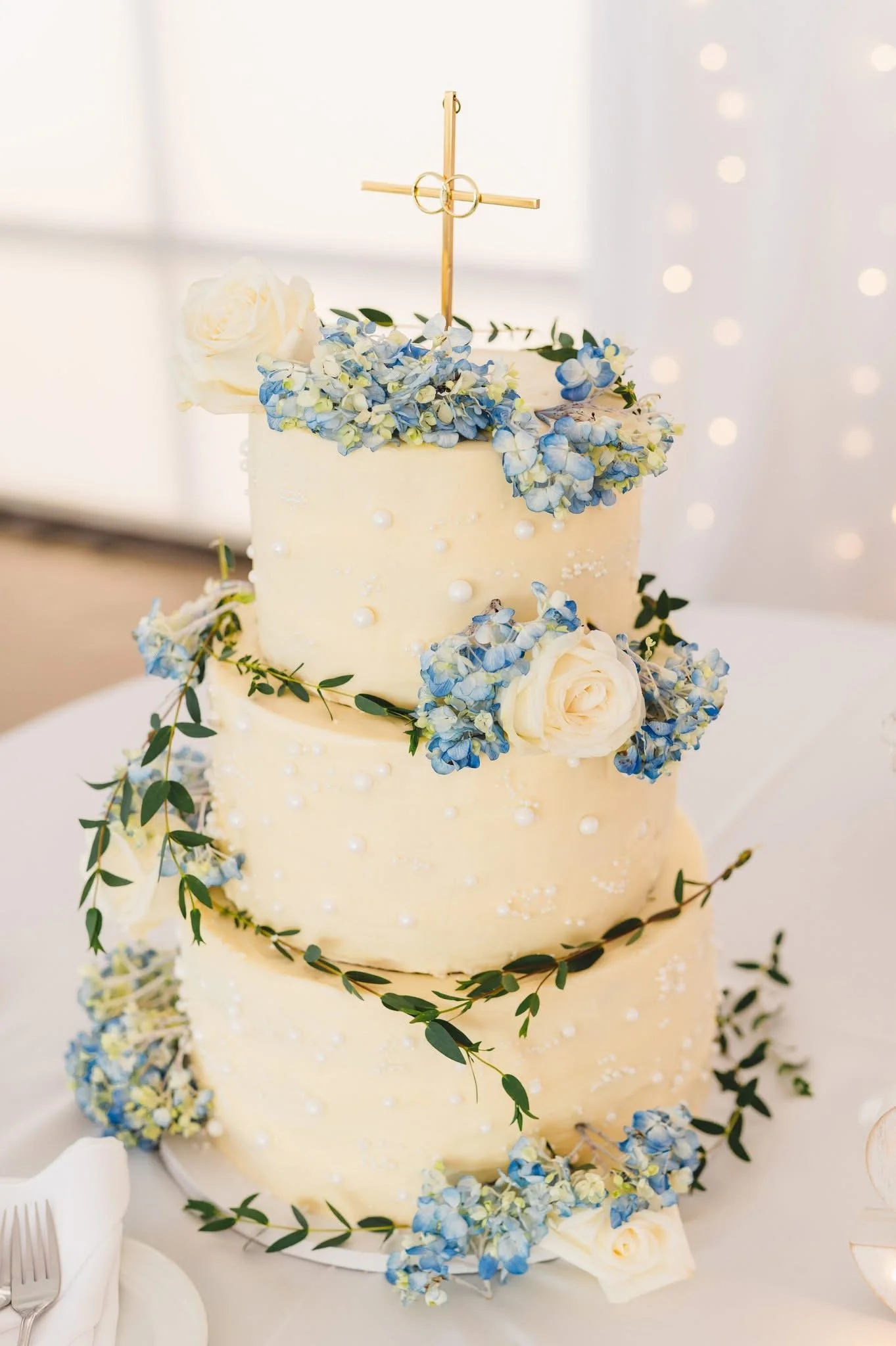 Three-tier white wedding cake decorated with white roses, blue and white hydrangeas, green foliage, and topped with a gold cross.