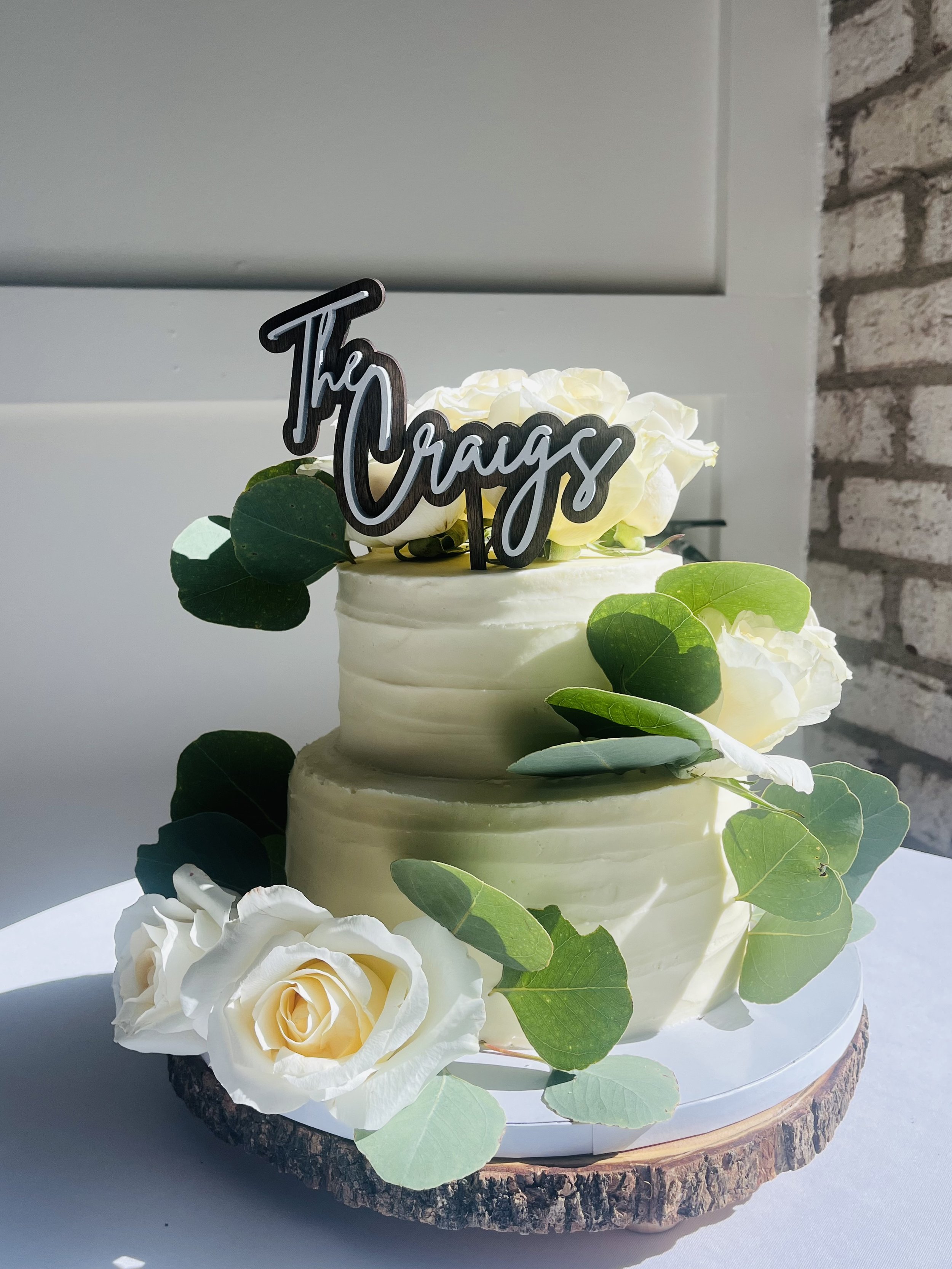 Two-tiered white wedding cake decorated with white roses and green leaves, with a black and white "The Crages" cake topper, placed on a wooden cake stand.