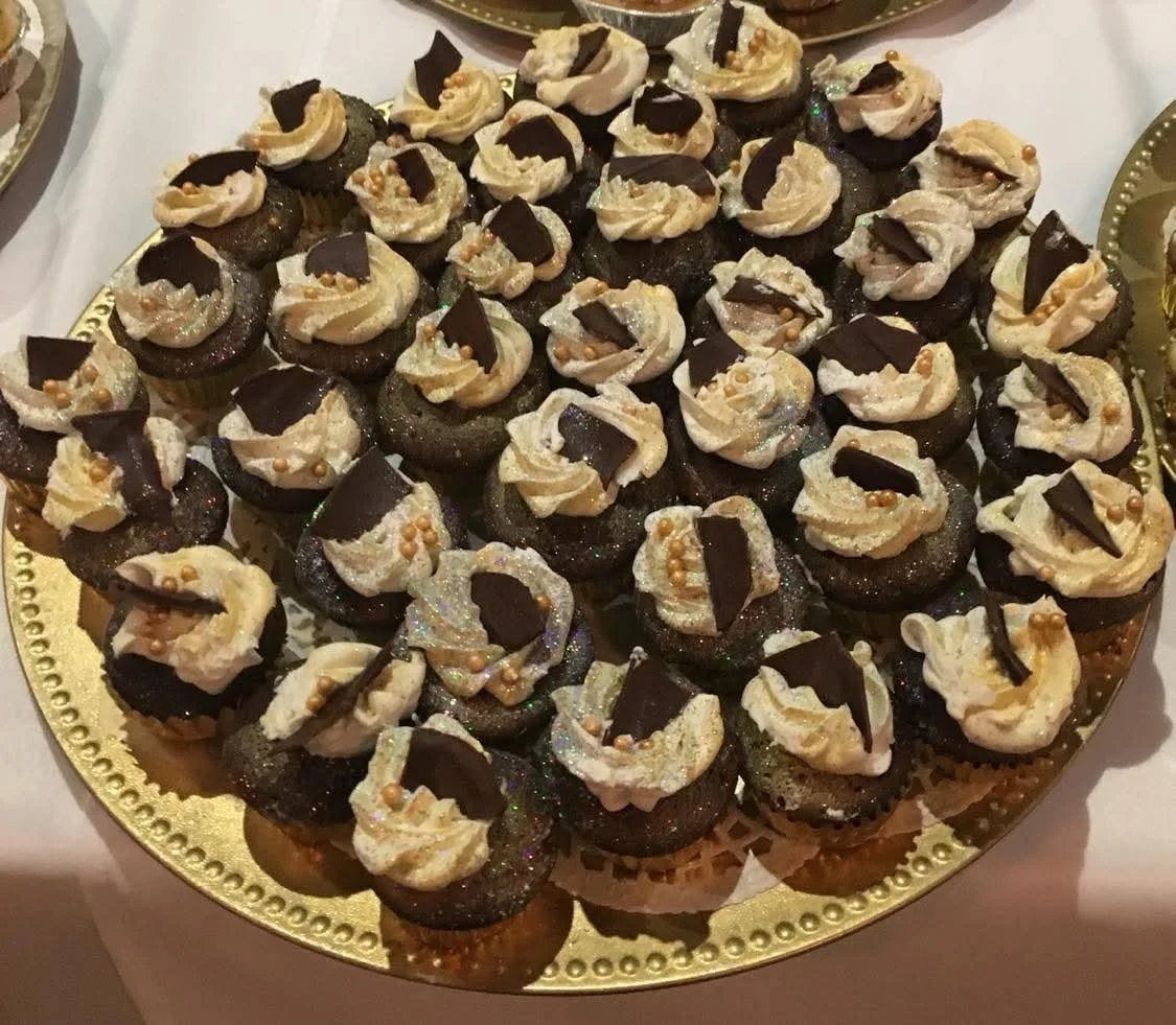 Plate of small chocolate cupcakes decorated with white frosting, gold edible beads, and a piece of dark chocolate on top.