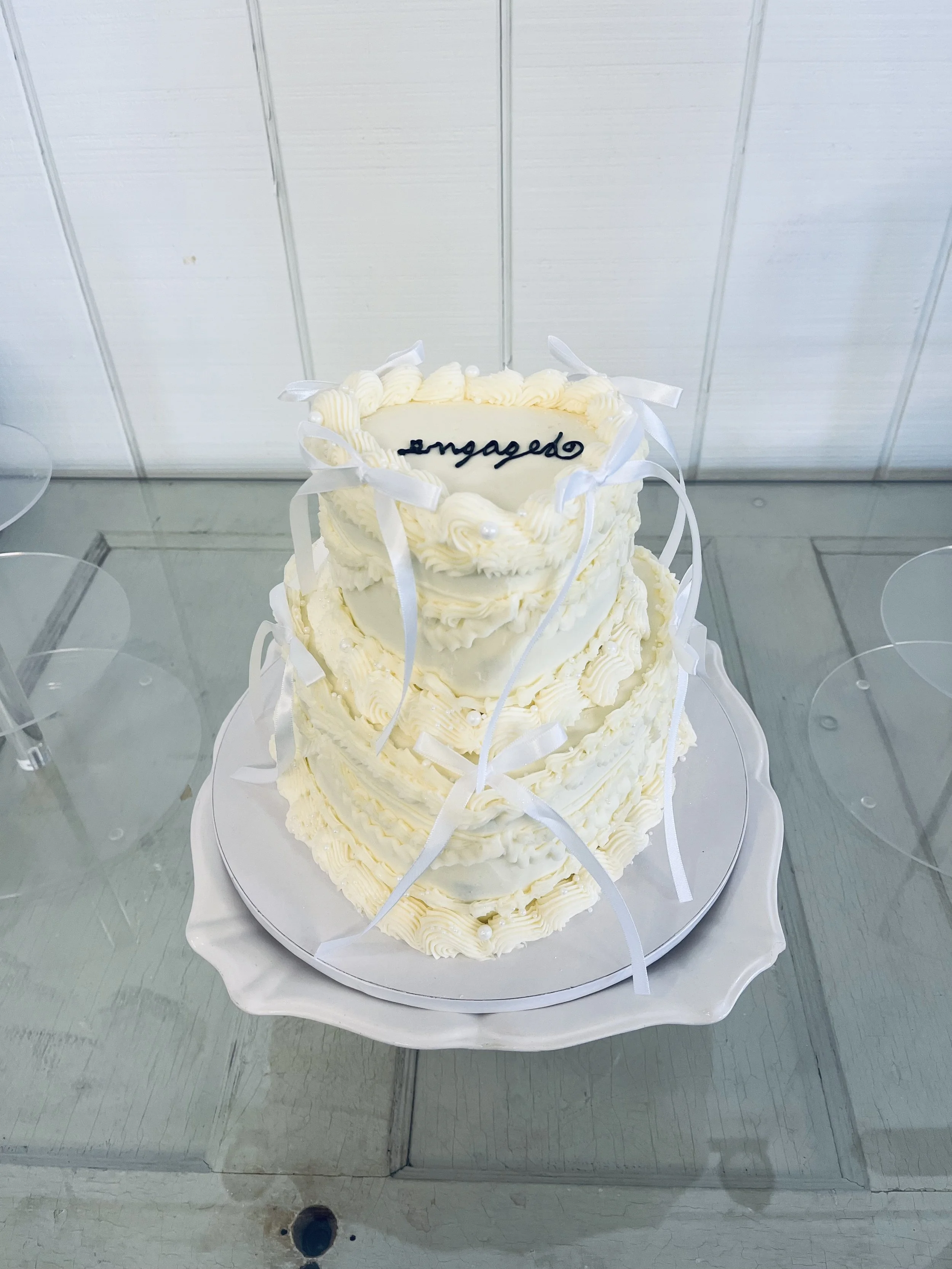 Three-tier wedding cake with white icing, ribbon decorations, and edible pearls, with a handwritten message that says 'engaged' on the top tier.