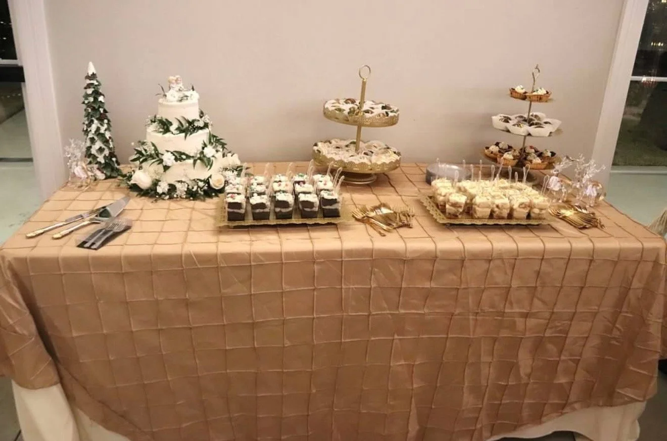 Dessert table with white cakes, cupcakes, and dessert trays, decorated with Christmas-themed decor and a gold-colored tablecloth.