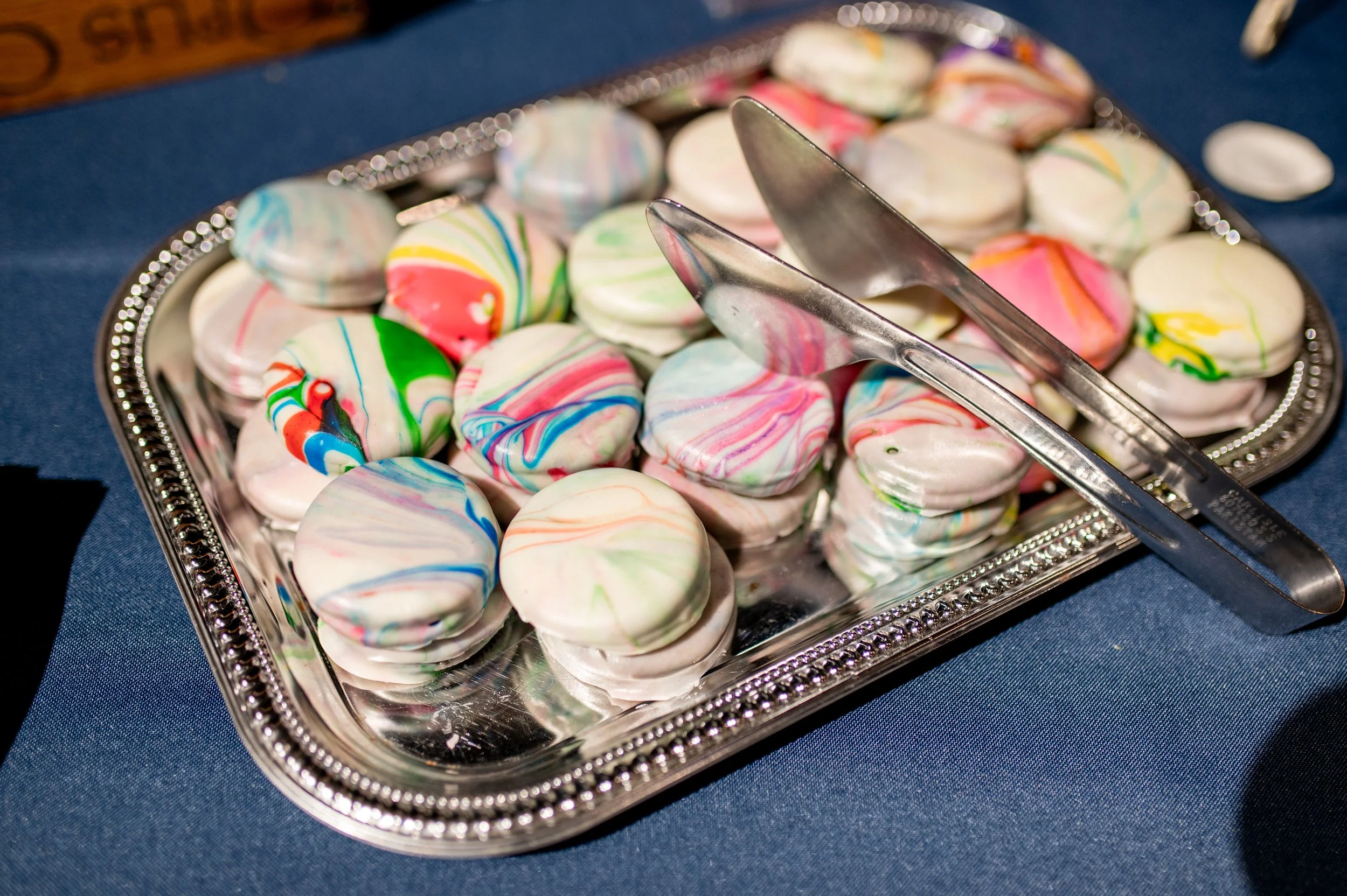 Colorful marbled small desserts on a silver serving tray with tongs, placed on a dark blue tablecloth.