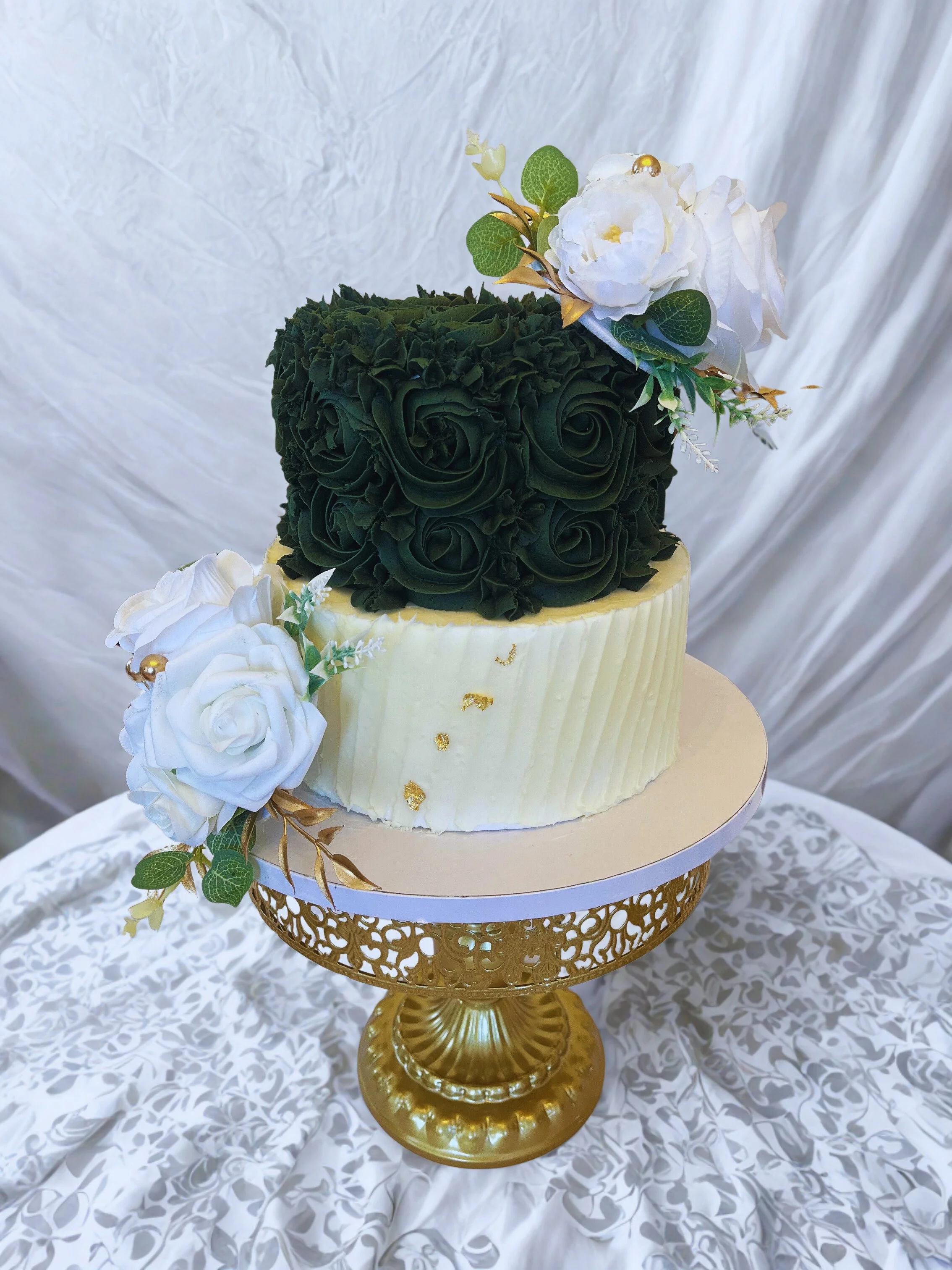 Two-tiered wedding cake with floral decorations on a gold cake stand, featuring white and dark green flower arrangements.