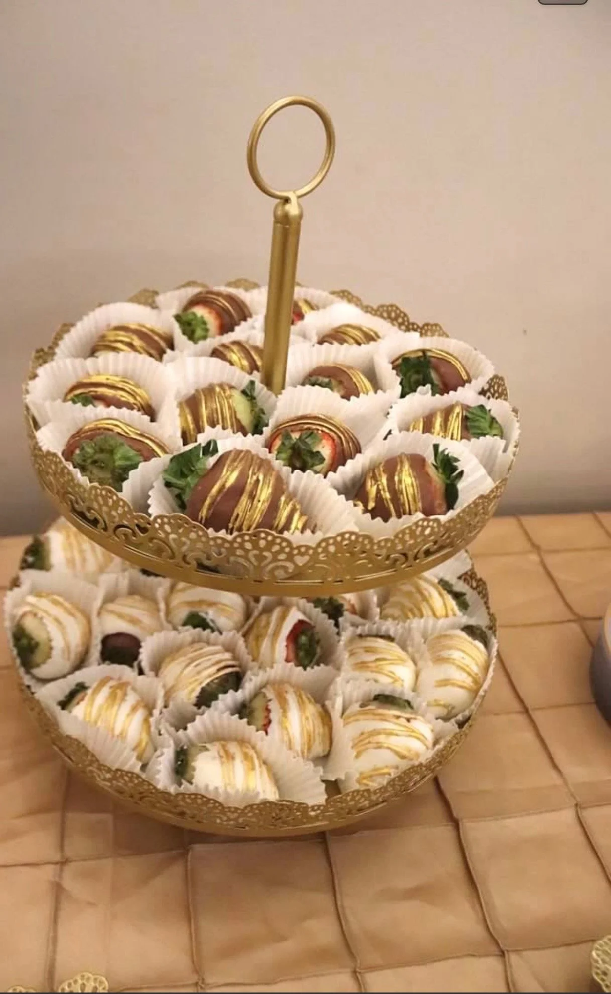 Two-tiered gold tray with assorted chocolate-covered strawberries decorated with gold drizzle and green leaves, arranged in white paper cups, placed on a tiled surface.