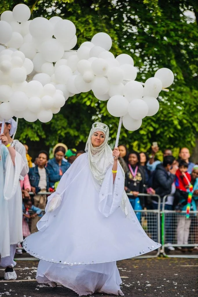 A woman dressed in white robe and headscarf holding a bunch of white balloons in a parade or celebration with a crowd of onlookers behind her.