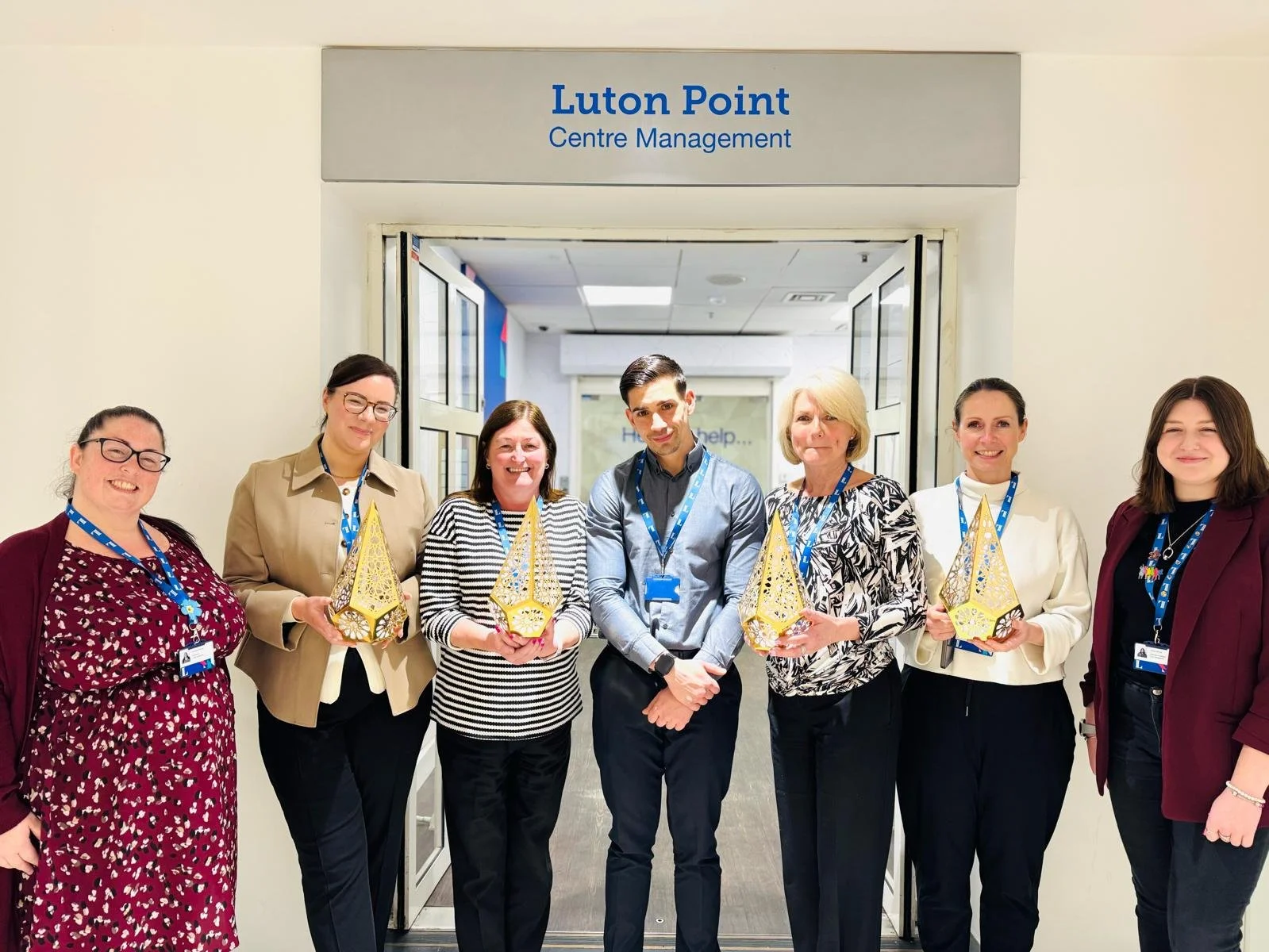 Group of seven people standing in front of a doorway with a sign that reads 'Luton Point Center Management', each holding a yellow decorative ornament.