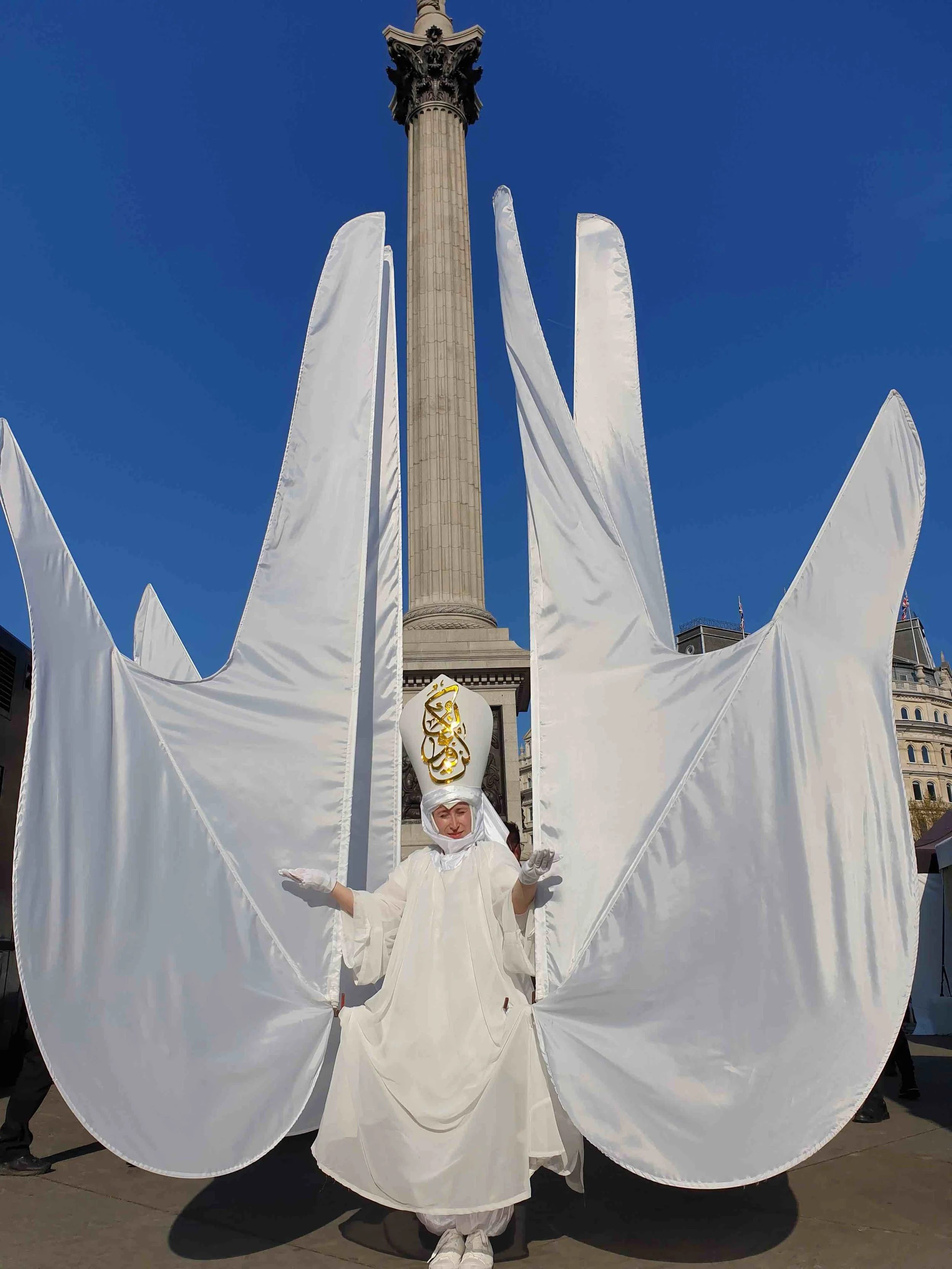 Person dressed in a white costume with large, wing-like fabric extensions on a city street during a parade or festival, with a tall monument in the background and a clear blue sky.