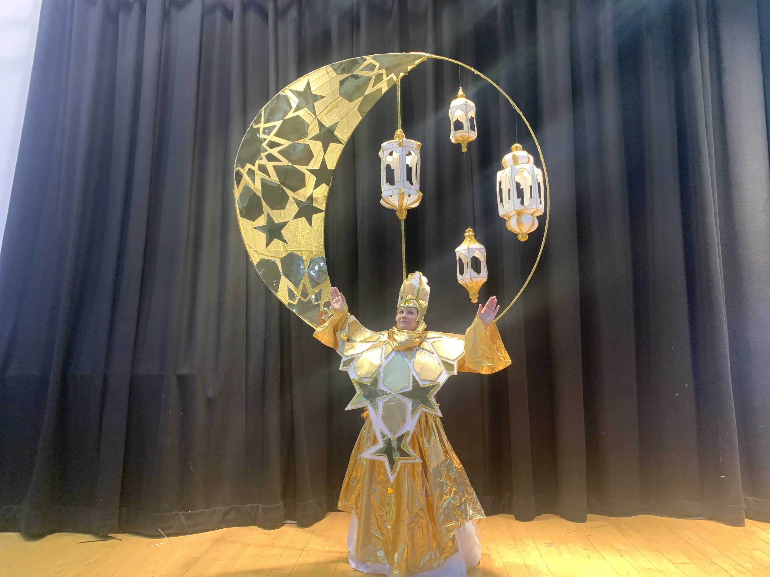 Person dressed in elaborate yellow and gold costume with star and moon decorations, standing on stage in front of black curtains, with lanterns hanging from a crescent moon structure.