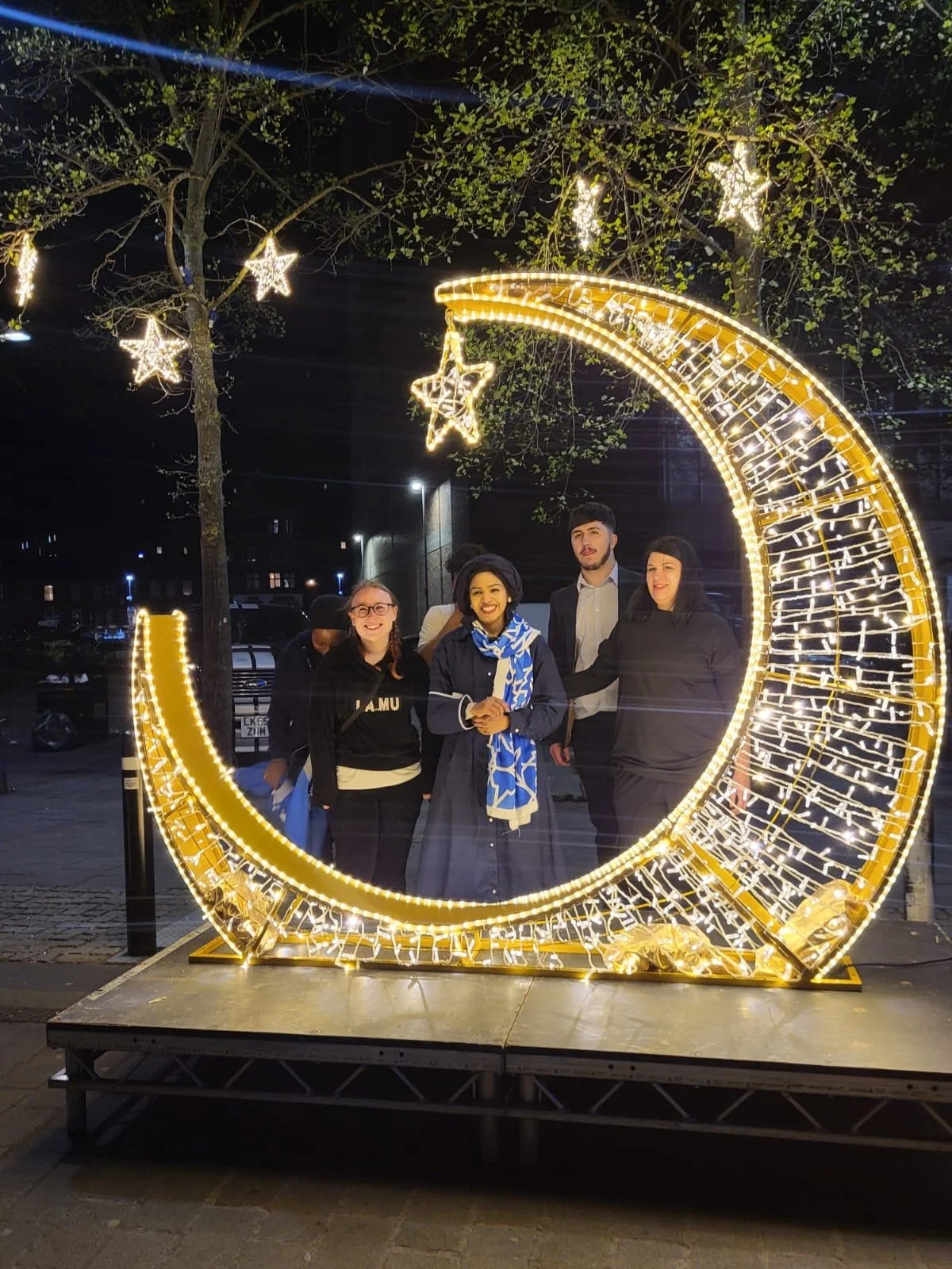 Group of five people standing inside a decorative crescent moon and star light display at night.
