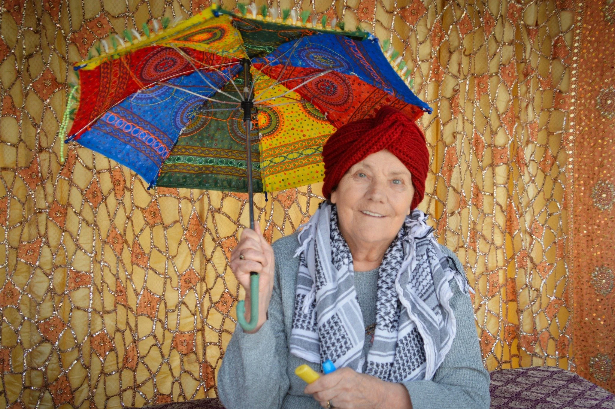 An elderly woman smiling, wearing a red knit headwrap, a grey sweater, and a black-and-white scarf, holding a colorful umbrella, with an ornate netted backdrop.