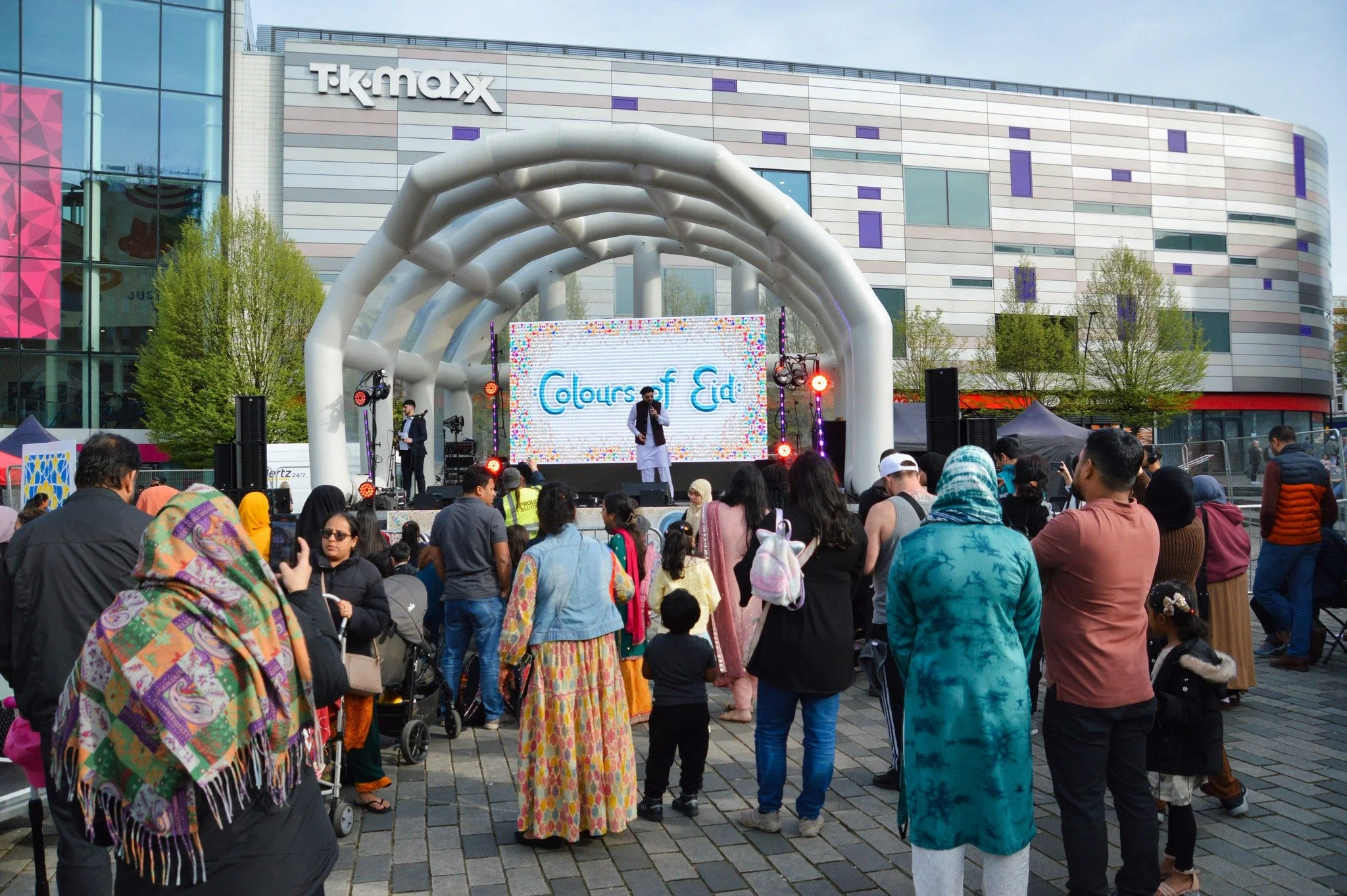 A crowd watching a stage performance at an outdoor event in front of a modern building with a TK Maxx sign. The stage has a large LED screen displaying 'Colors of Eid,' and performers are on stage with colorful lighting.