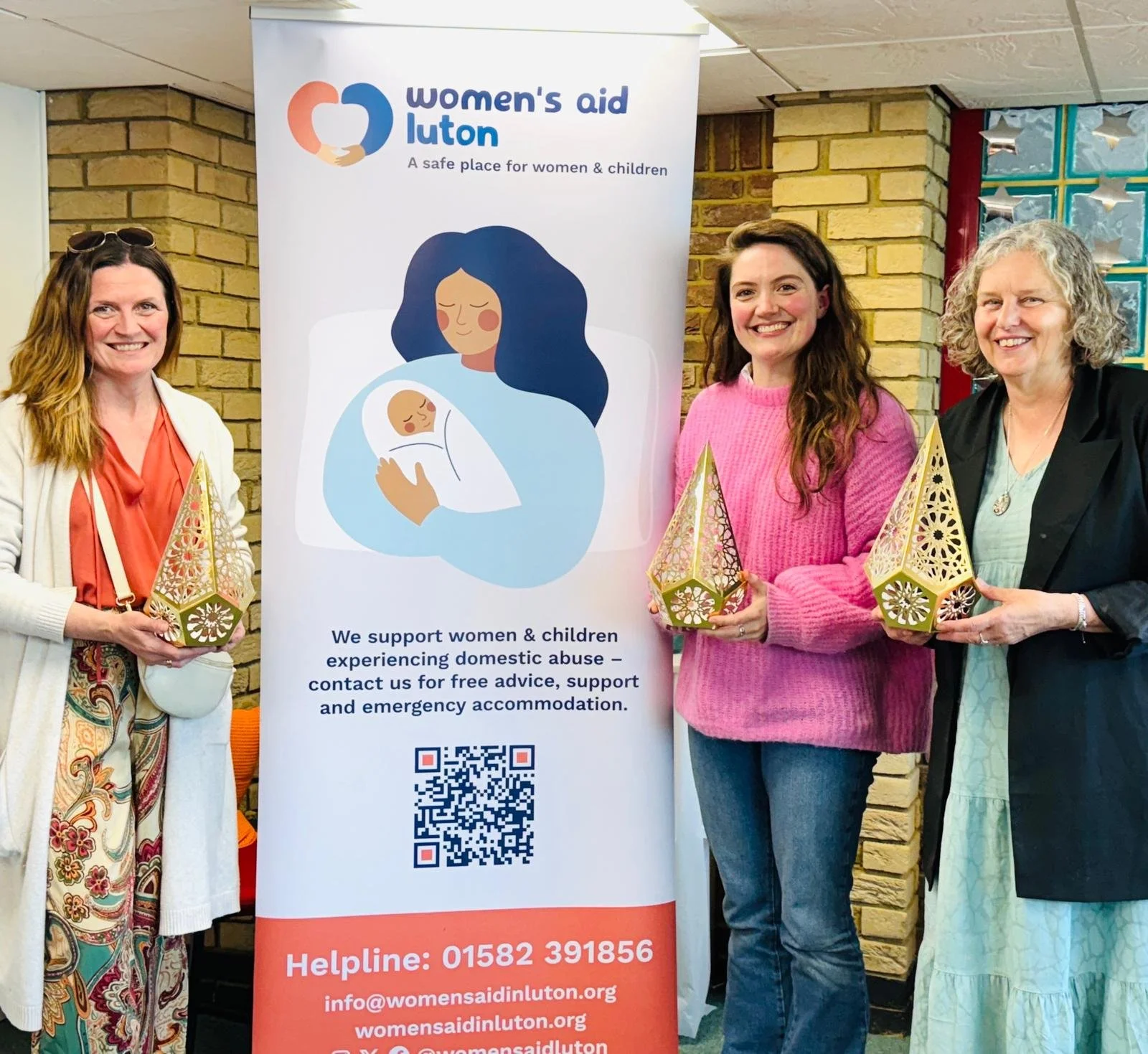 Three women standing in front of a Women's Aid Luton banner, each holding a decorative gold and pink lantern. The banner displays illustrations of a woman holding a baby and text about supporting women and children experiencing domestic abuse.