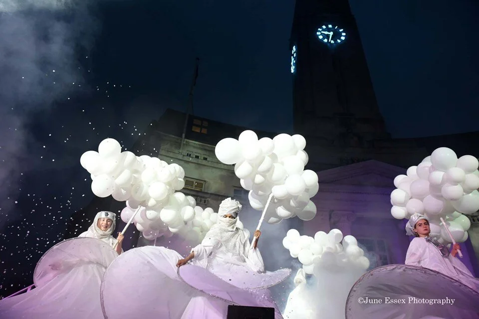 Three women dressed in white costumes with large skirts, holding bunches of white balloons, part of a parade or festival at night with a clock tower in the background.