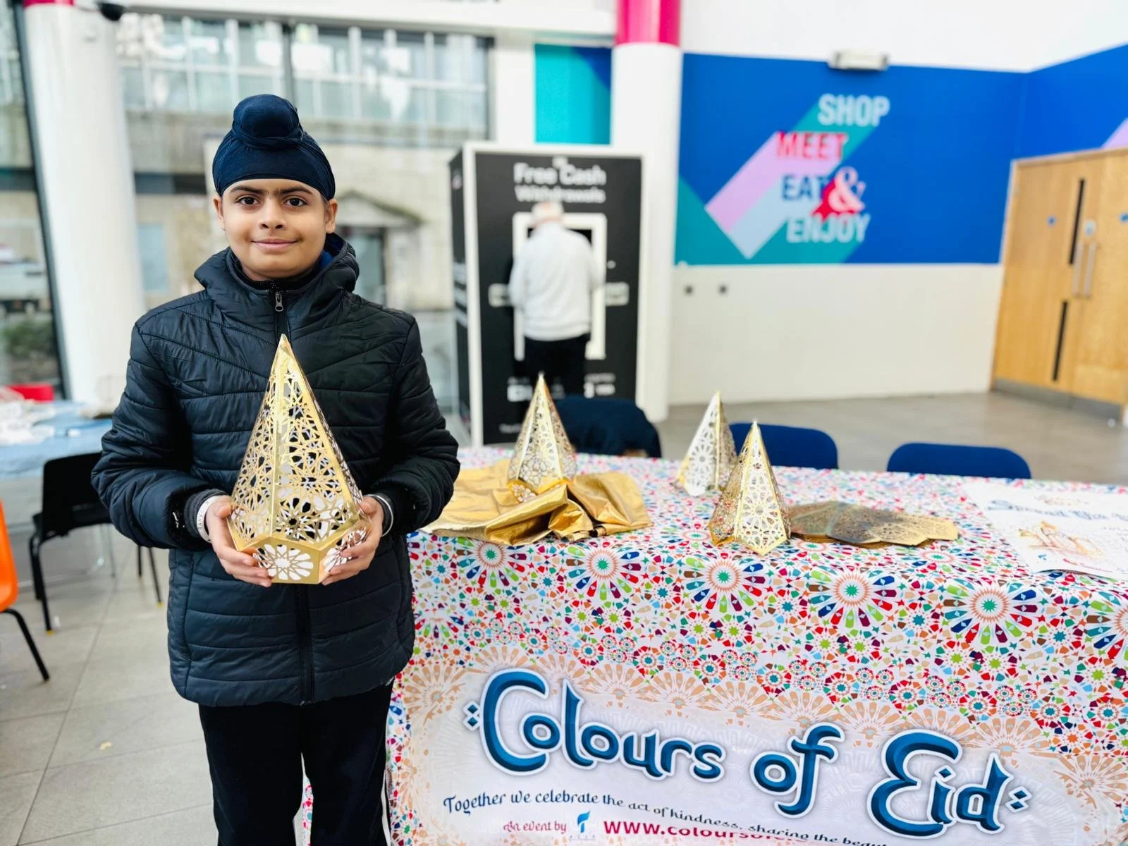 A boy holding a decorative gold paper lantern at an event called 'Colors of Eid' with a colorful patterned tablecloth and matching lanterns on the table.