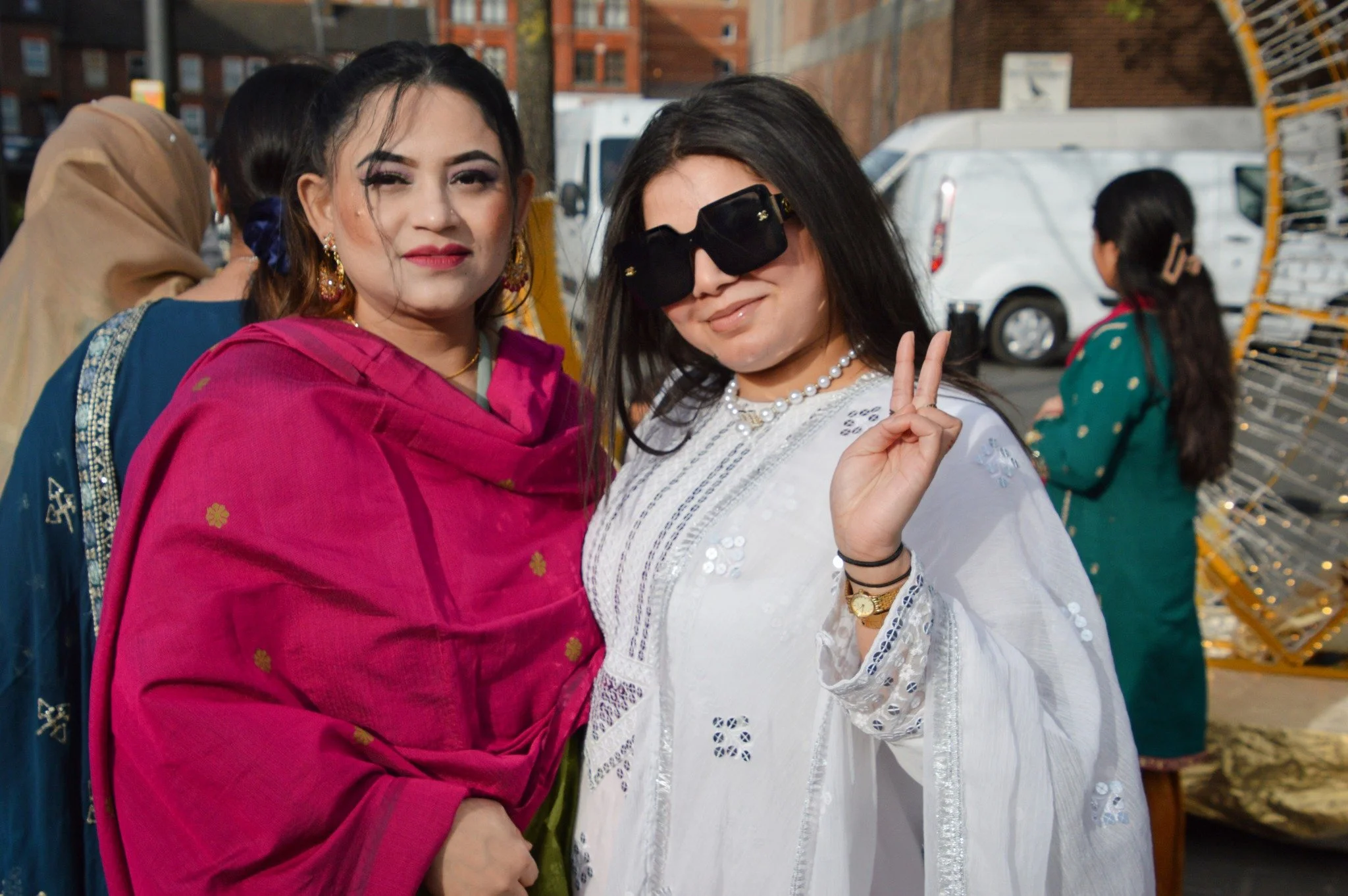 Two women posing outdoors: one in a pink traditional outfit with earrings and makeup, the other in a white outfit with sunglasses and a pearl necklace, making a peace sign.