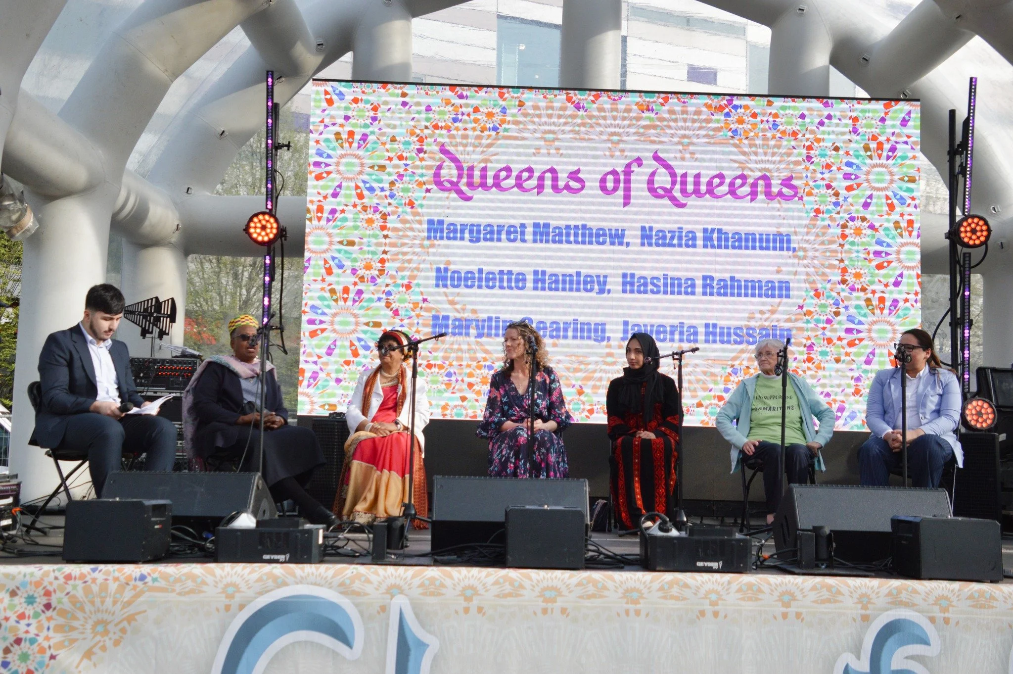 Six women and one man sitting on stage at an outdoor event, with a colorful digital screen in the background displaying names of women recognized as queens.