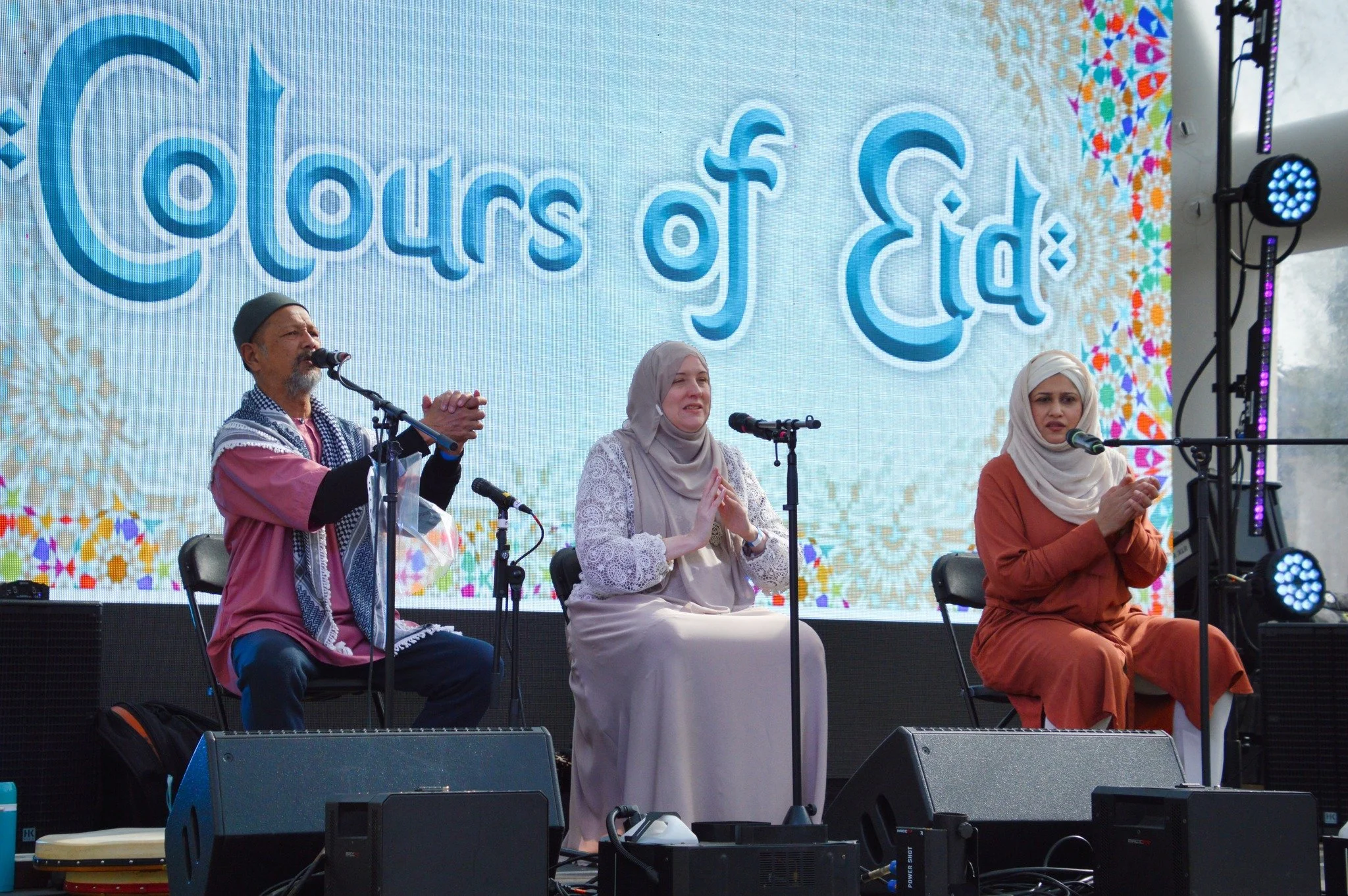 Three people sitting on chairs on a stage with a large screen behind them displaying the text 'Colors of Eid'. They are participating in a cultural or religious event, with microphones in front of them. The man on the left is wearing a pink shirt and