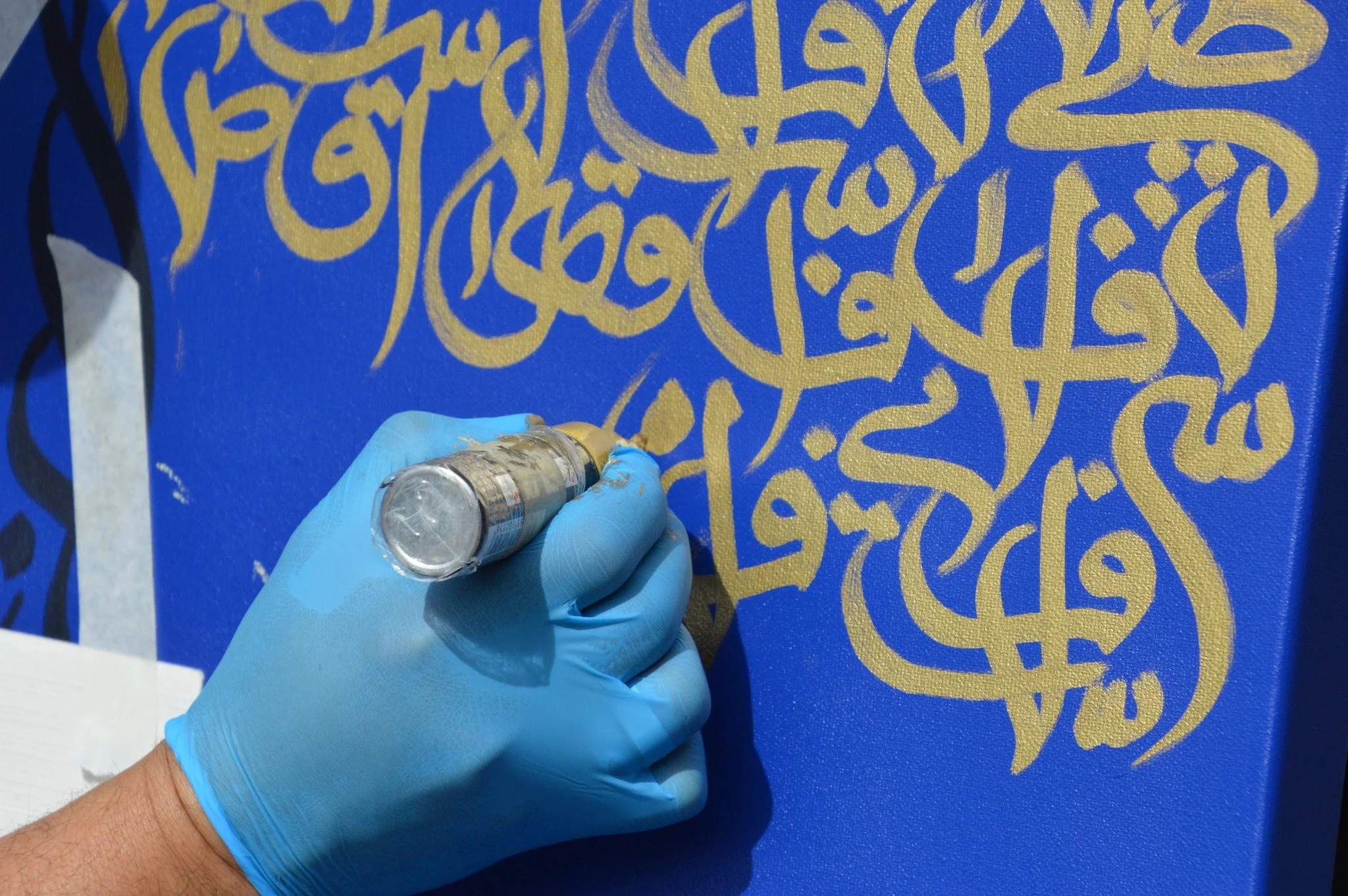 A person wearing a blue glove is creating gold Arabic calligraphy on a blue canvas using a spray paint can.