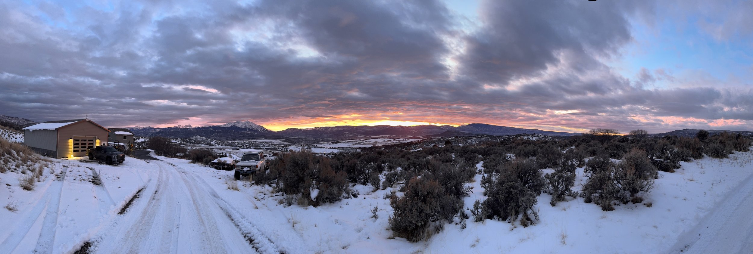 Snow-covered landscape at sunset with mountains in the distance, a garage with cars nearby, and cloudy sky with hints of pink and orange.