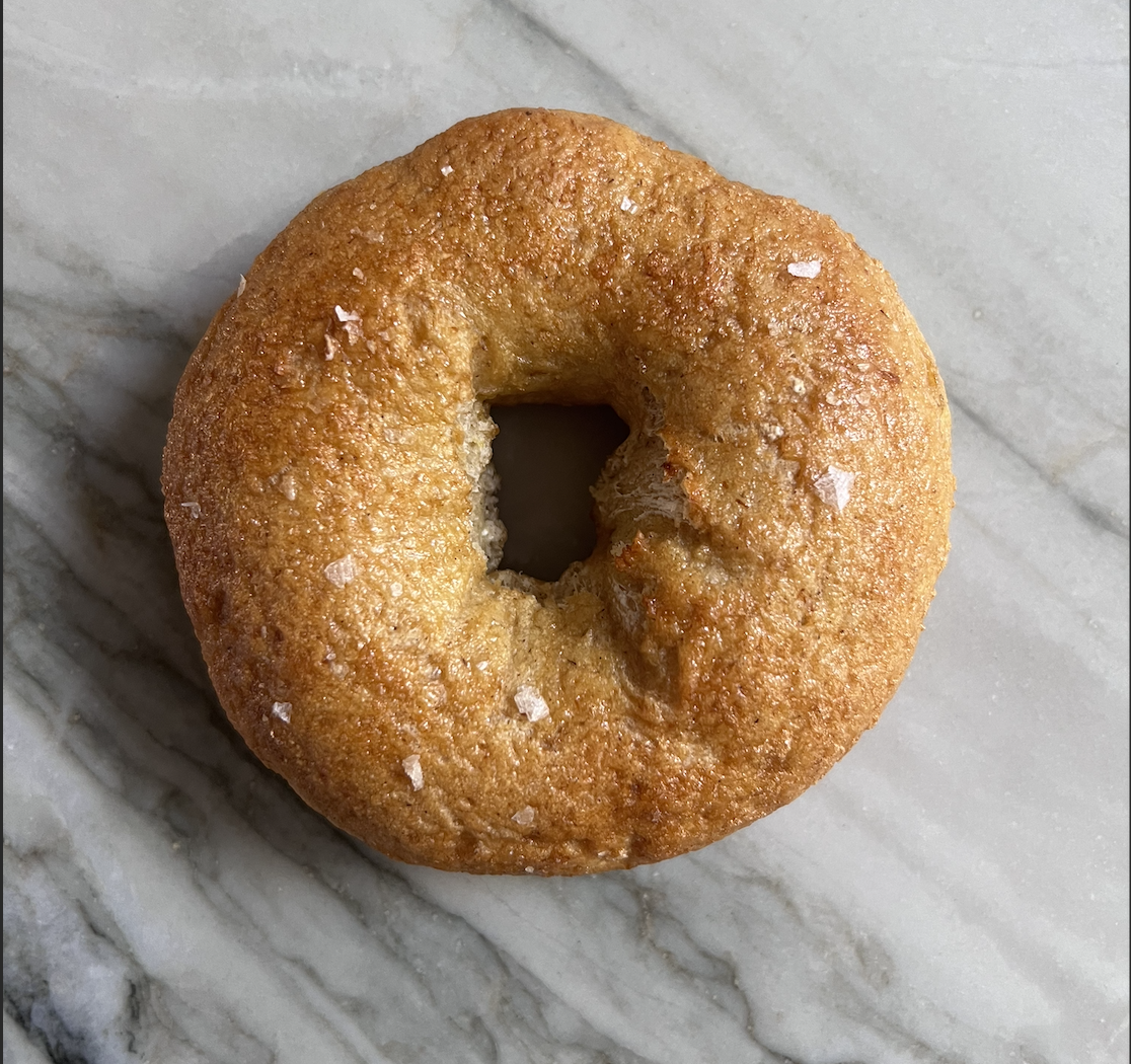Close-up of a freshly baked, golden brown, circular donut with a small hole in the center, topped with coarse sea salt, placed on a marble surface.