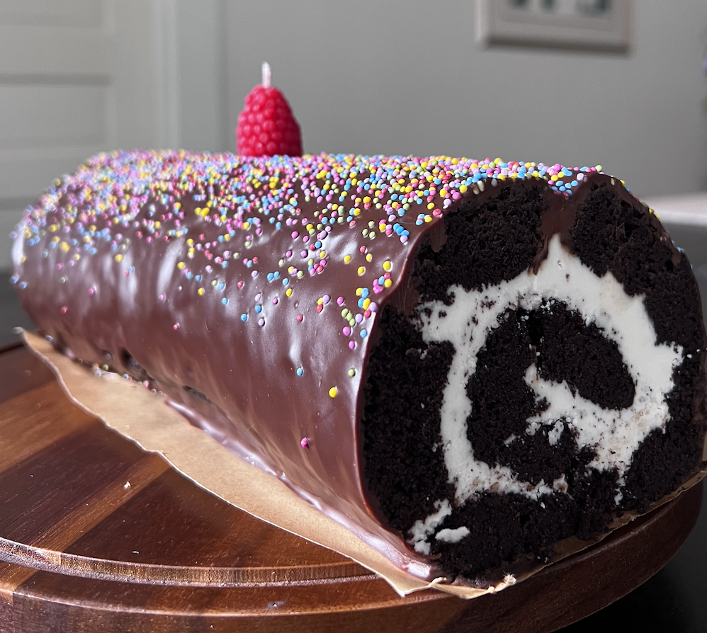 Chocolate roll cake with colorful sprinkles and a raspberry on top, placed on a wooden board.