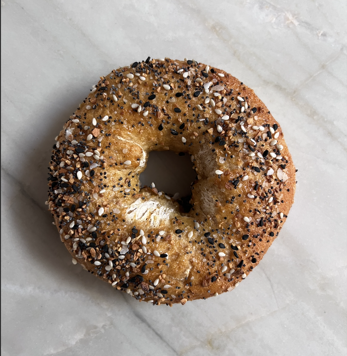 Close-up of a bagel topped with mixed seeds and spices, resting on a light-colored marble surface.