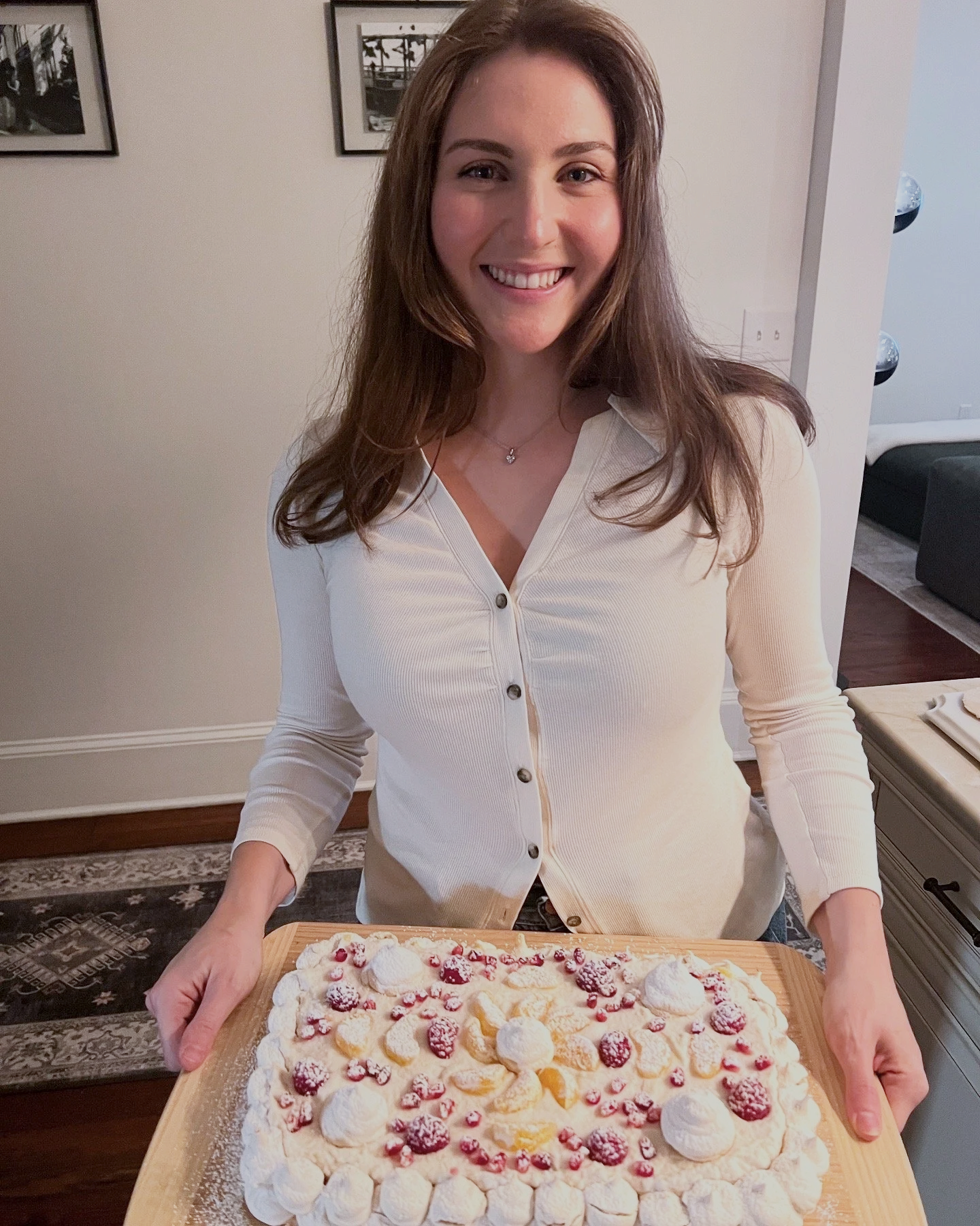 Woman holding a large tray of decorated dessert with whipped cream, berries, and fruit slices, smiling in a home setting.